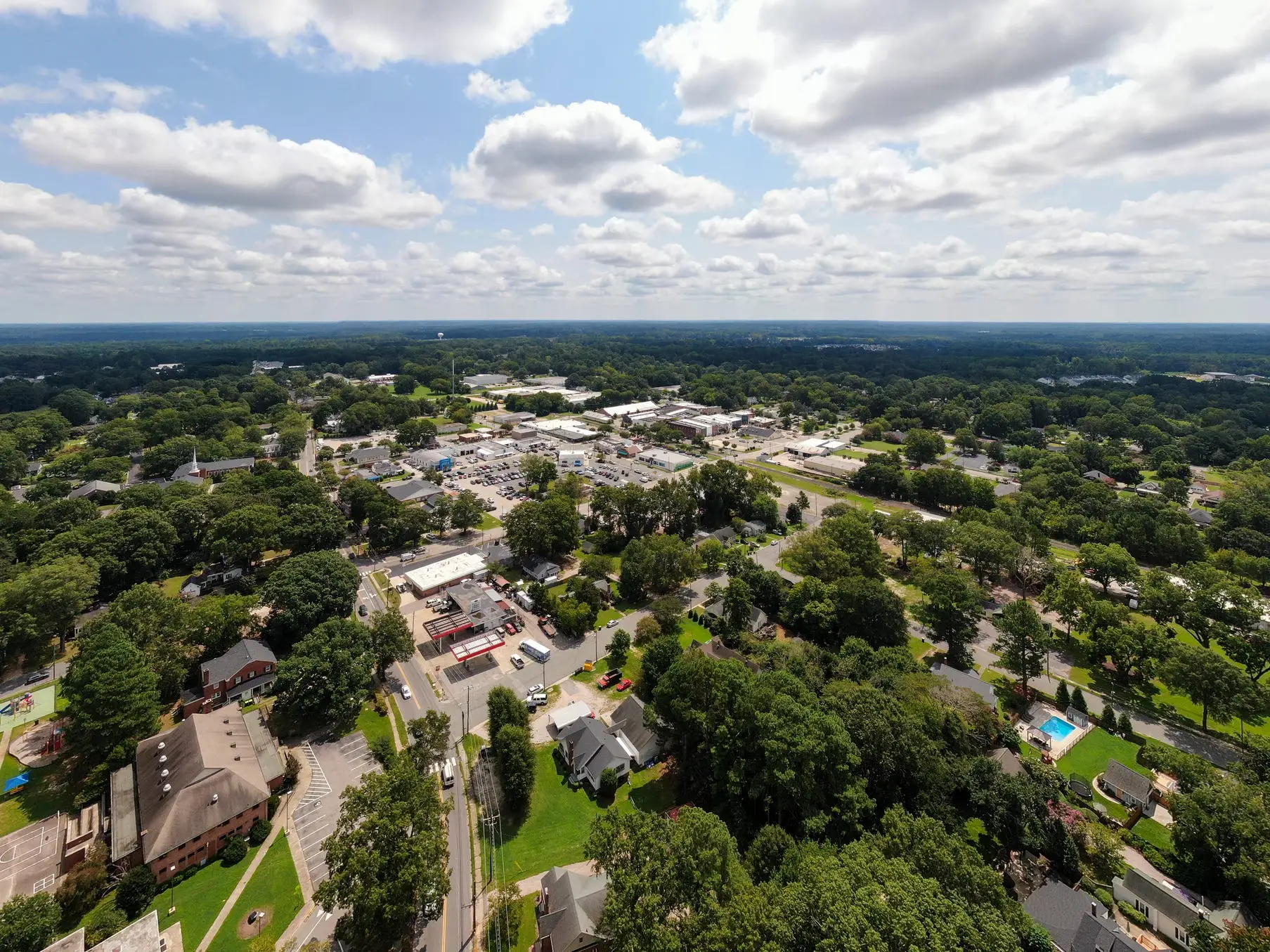 Aerial view of a leafy suburban town with roads, low buildings, homes, and a partly cloudy sky.