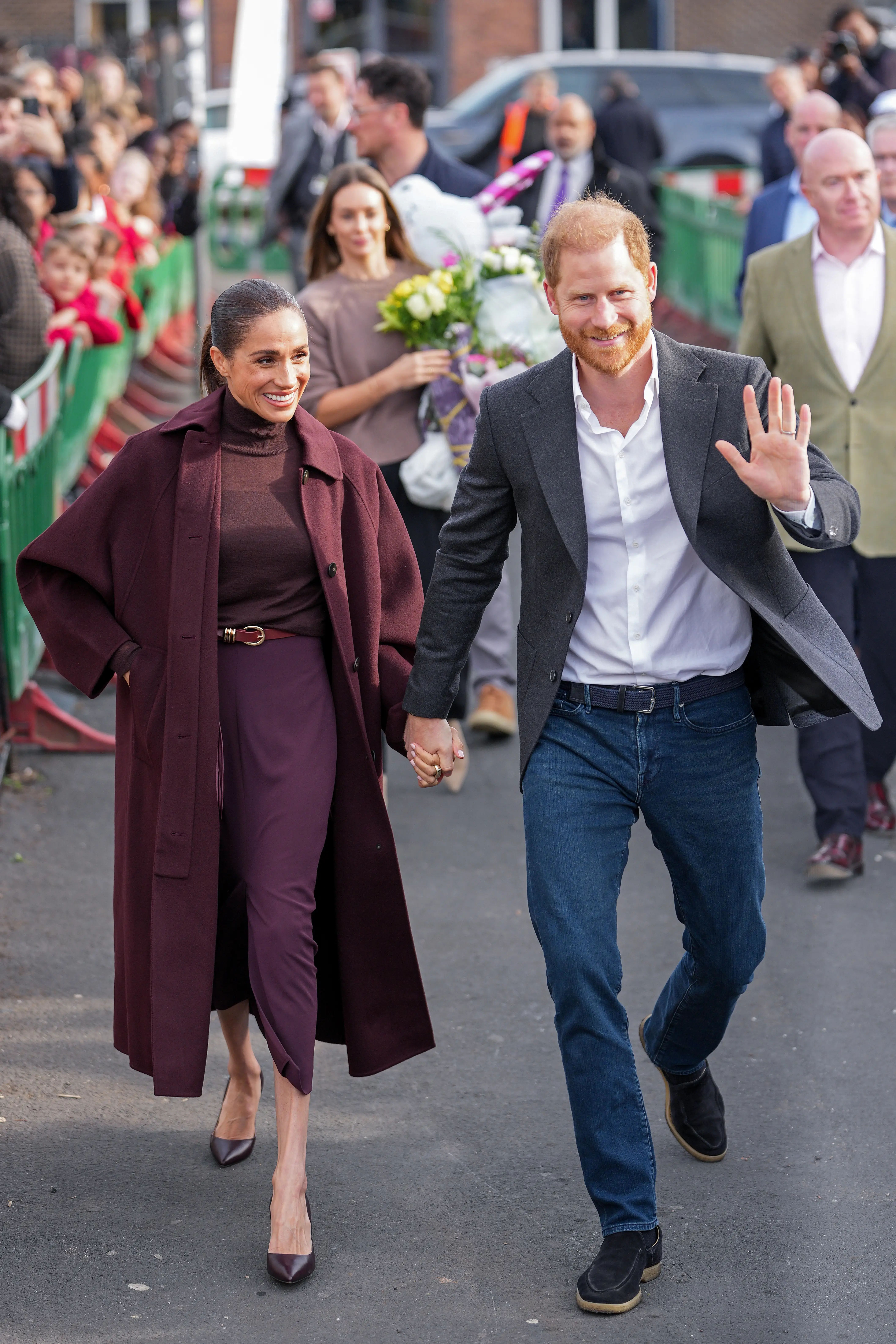 Meghan Markle and Prince Harry walk hand in hand past a crowd, with Harry waving.