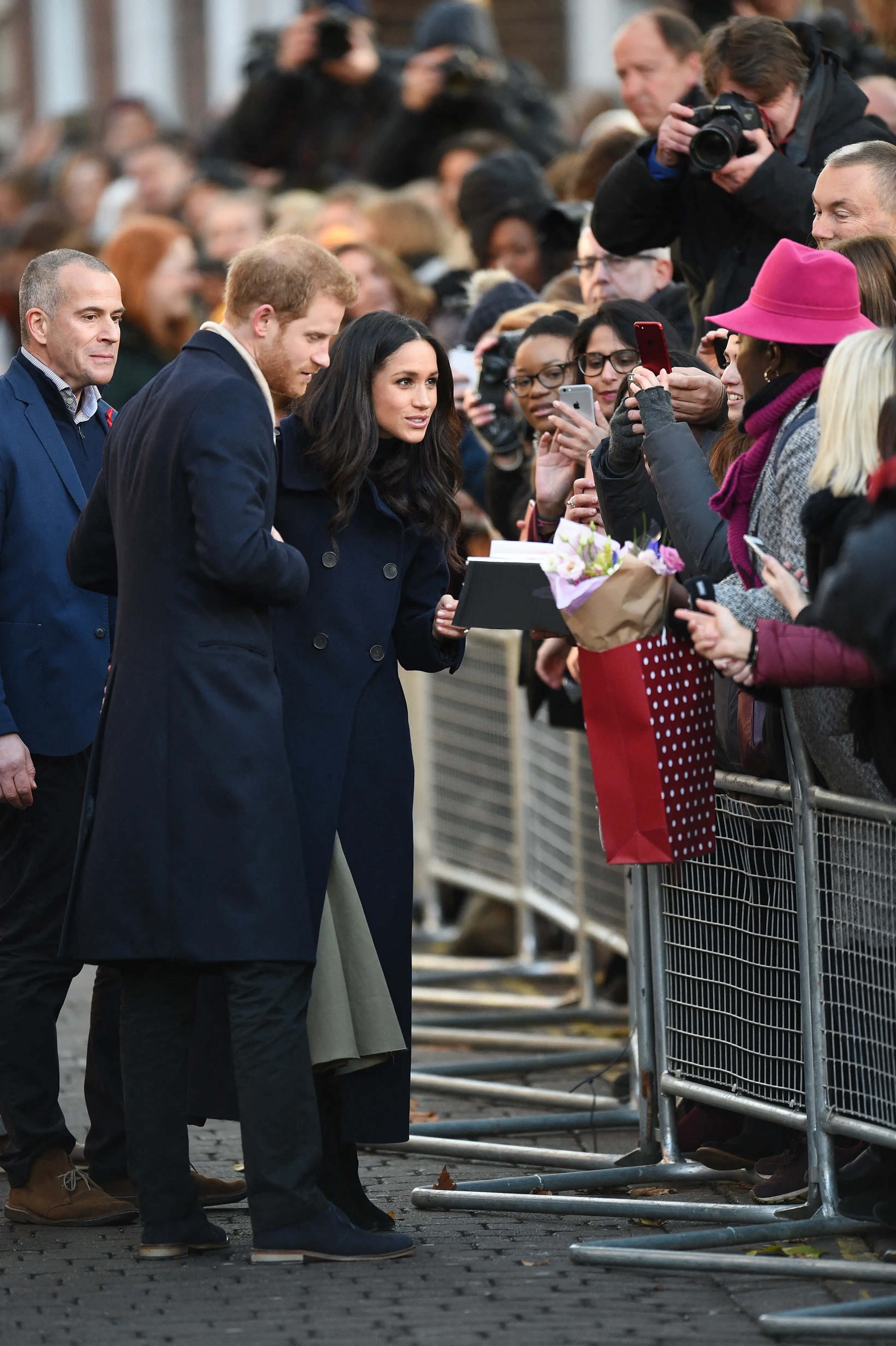 Prince Harry and Meghan Markle greet a crowd behind metal barricades while people take photos and hold flowers.