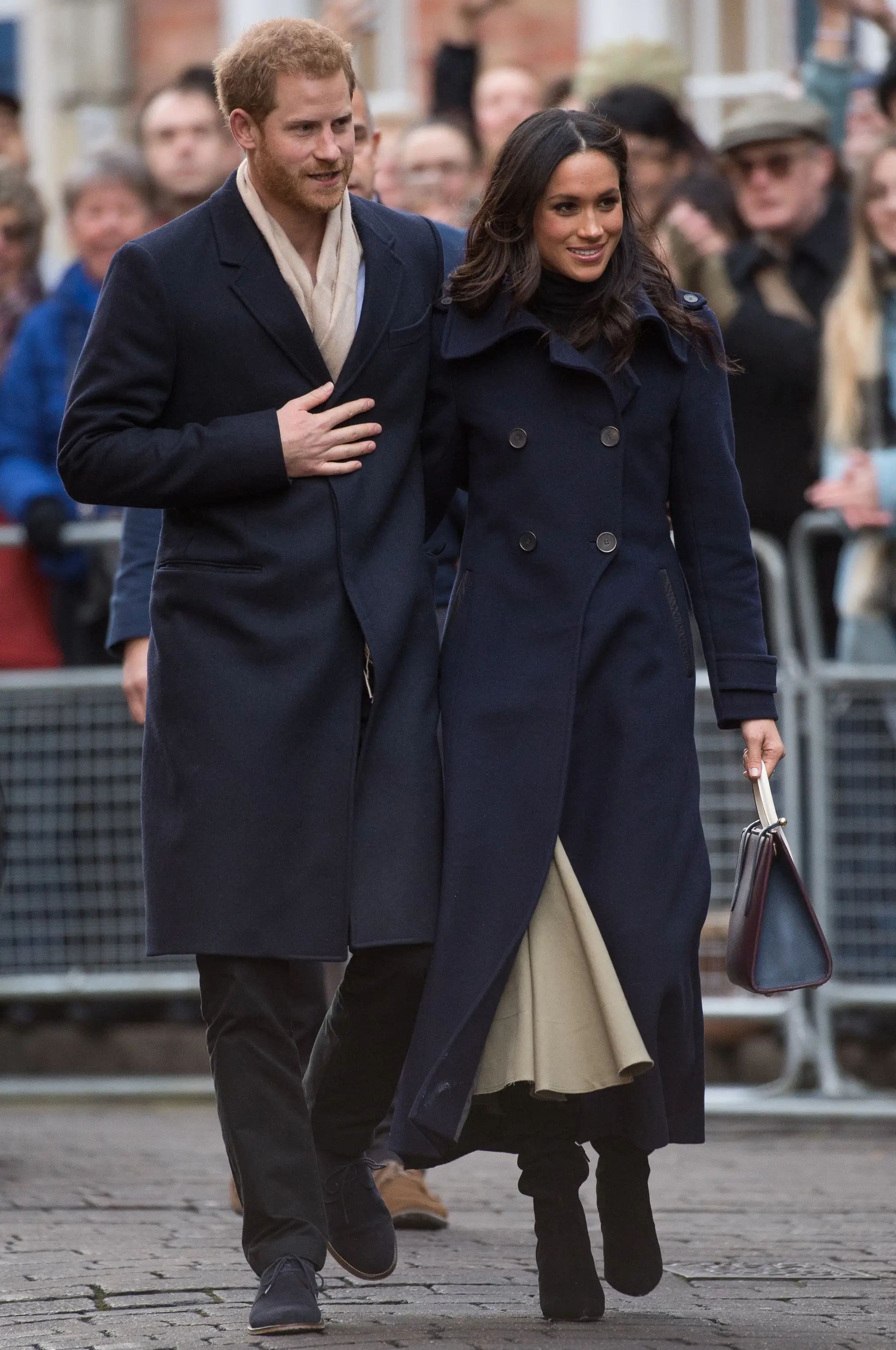 Prince Harry and Meghan Markle walk together past a crowd, wearing dark coats on a paved street.