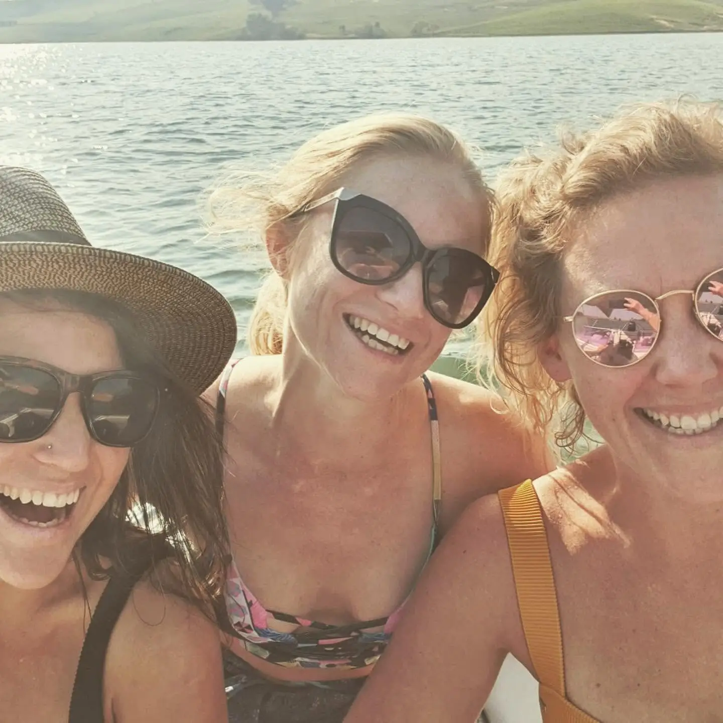 Three people wearing sunglasses smile in a sunny selfie on water with hills in the background.