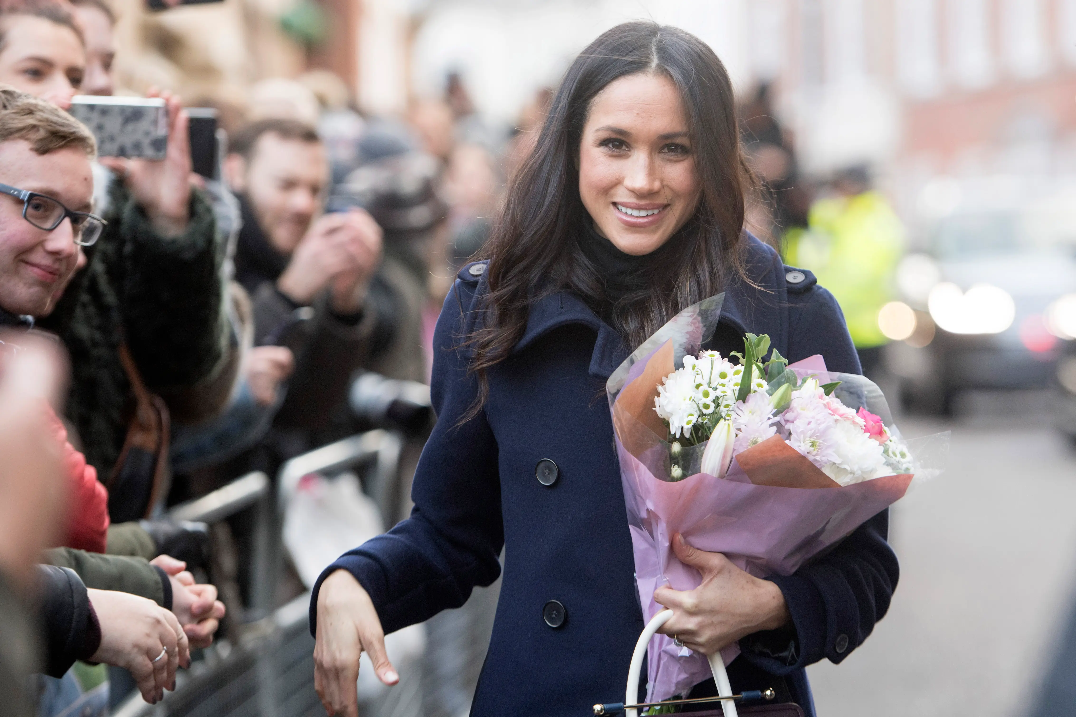 Meghan Markle smiles while holding a bouquet of flowers beside a crowd and photographers on a city street.