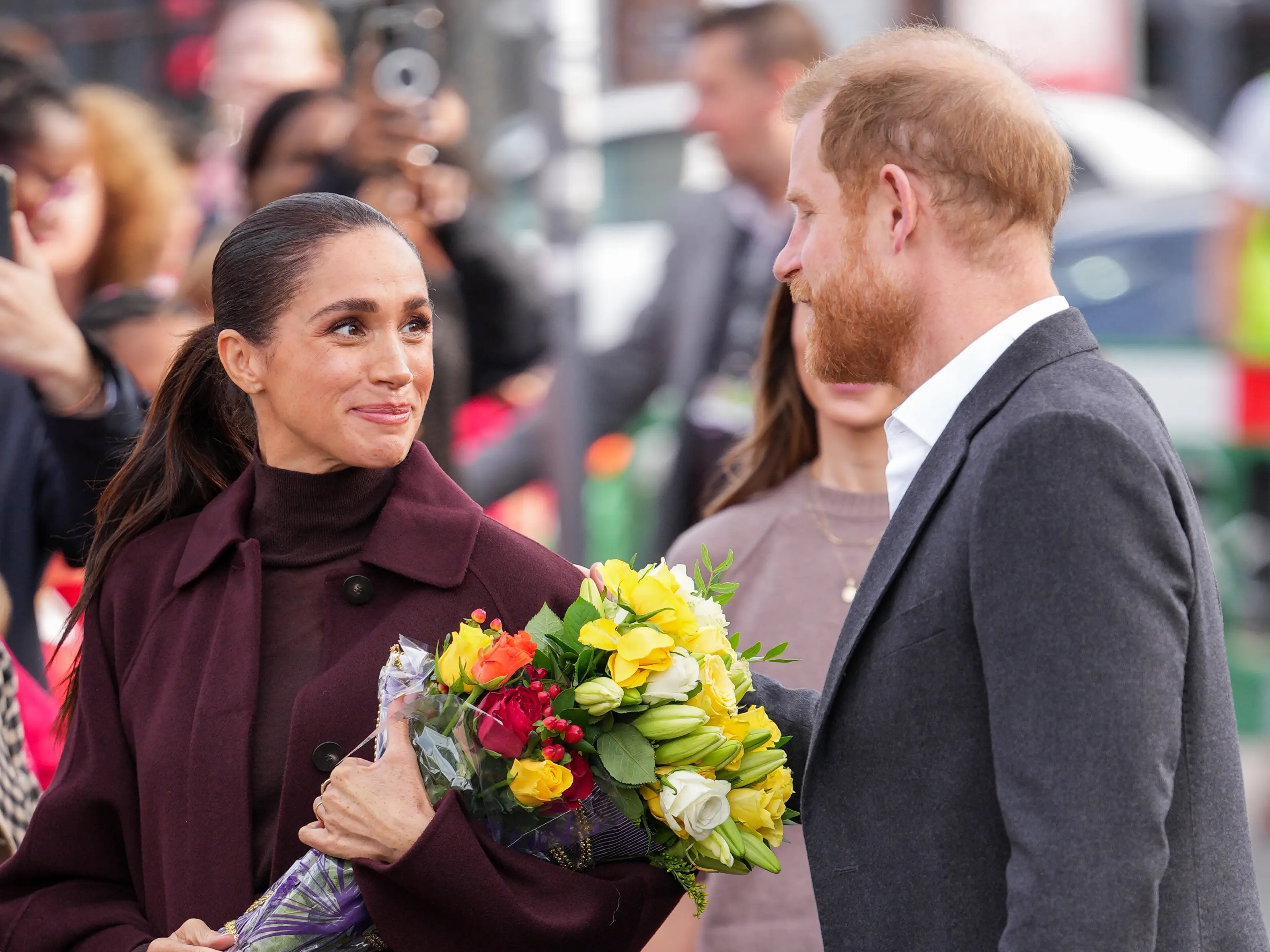 Meghan Markle holds flower bouquets while standing beside Prince Harry during a public outdoor appearance.