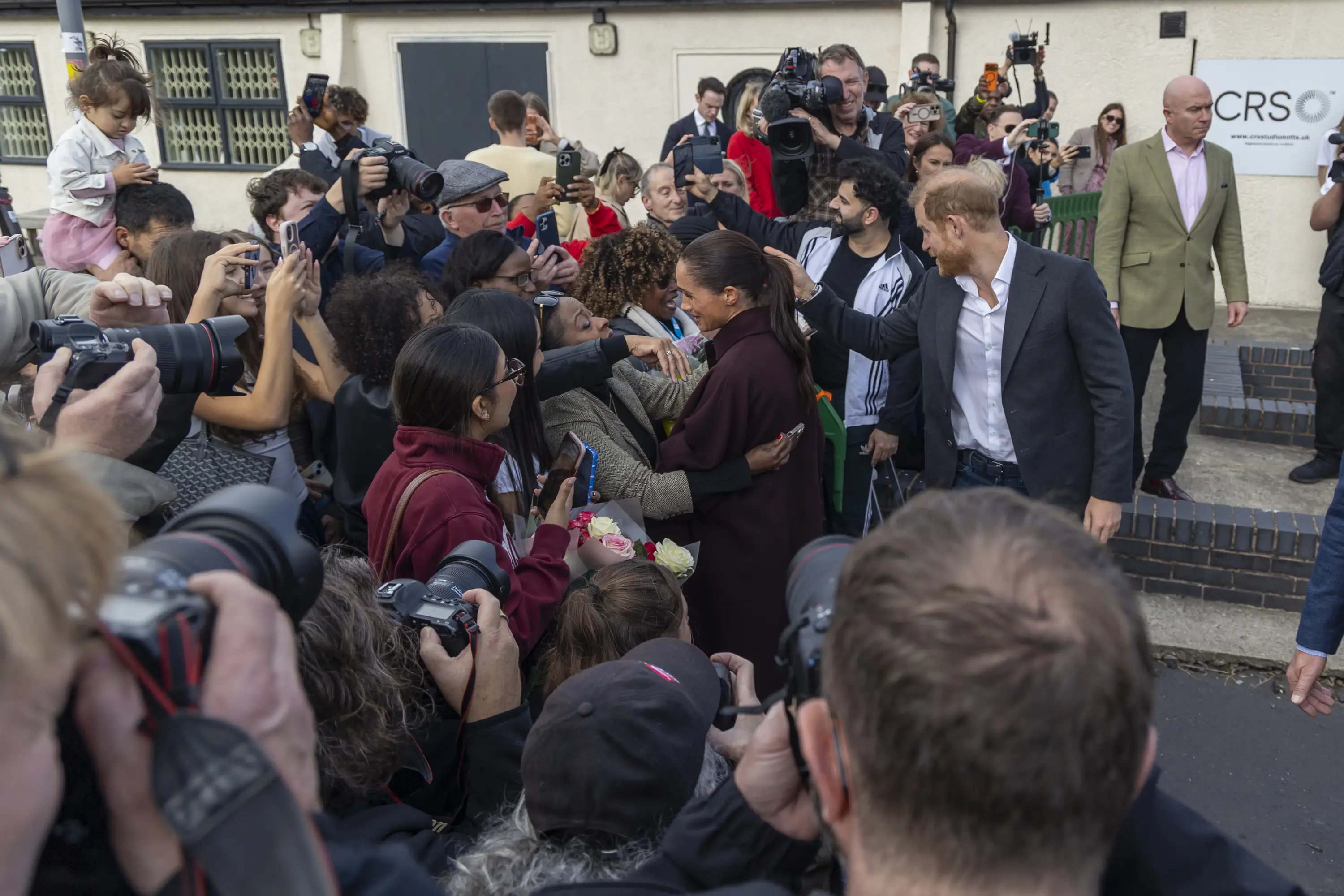 Meghan, Duchess of Sussex, greets a crowd beside Prince Harry as photographers gather outside a building.