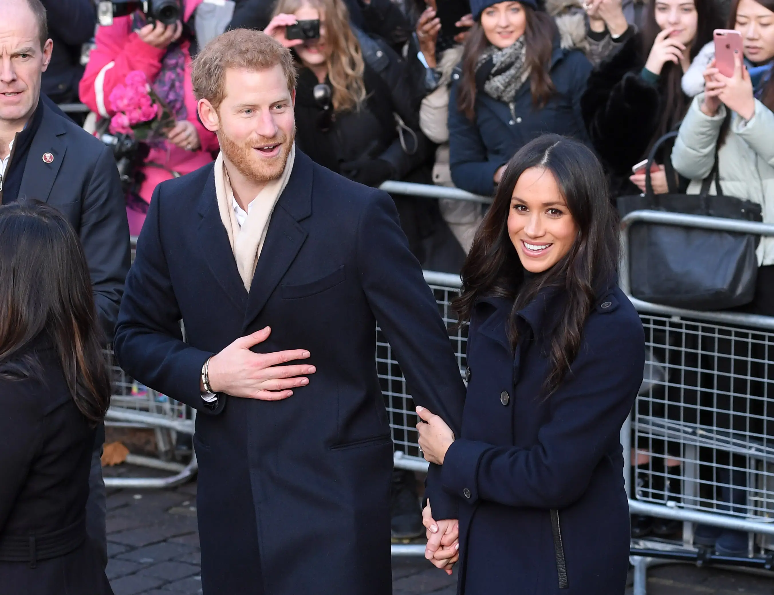 Prince Harry and Meghan Markle walk hand in hand beside crowd barriers as onlookers take photos.