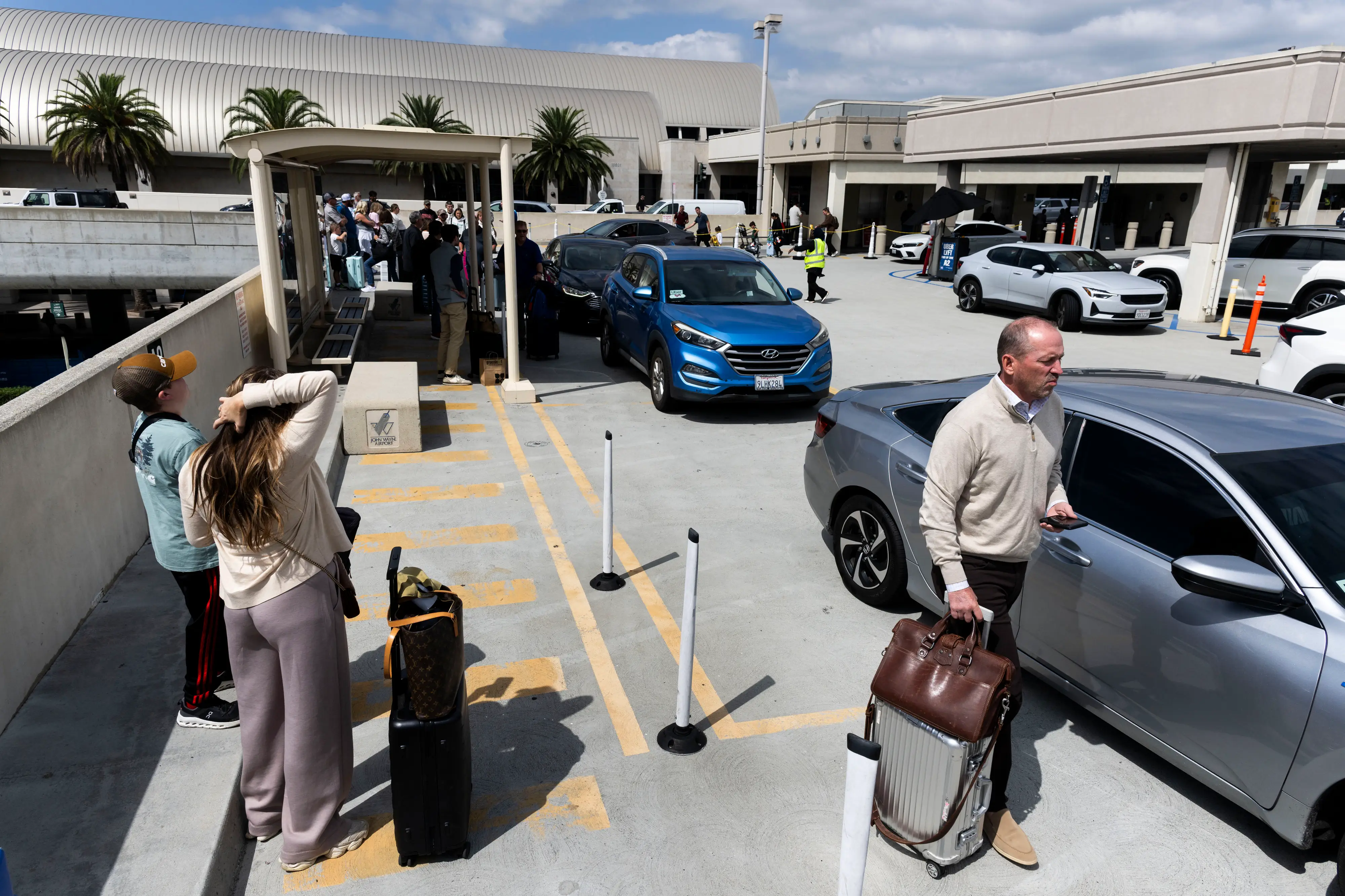 Travelers with luggage wait near cars at a busy terminal curbside pickup area under partly cloudy skies.