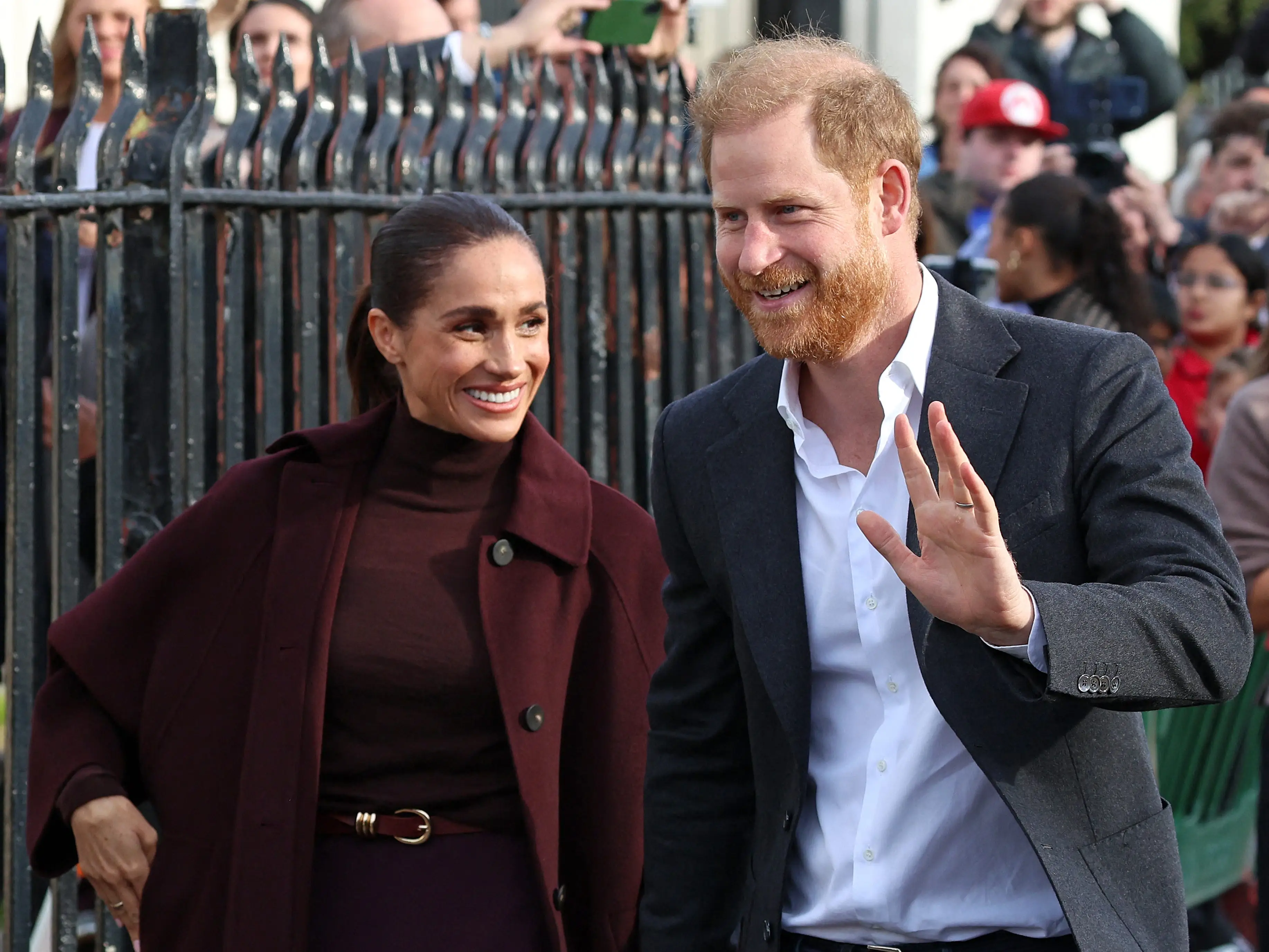 Meghan Markle and Prince Harry hold hands as they walk past a crowd, with Prince Harry waving.
