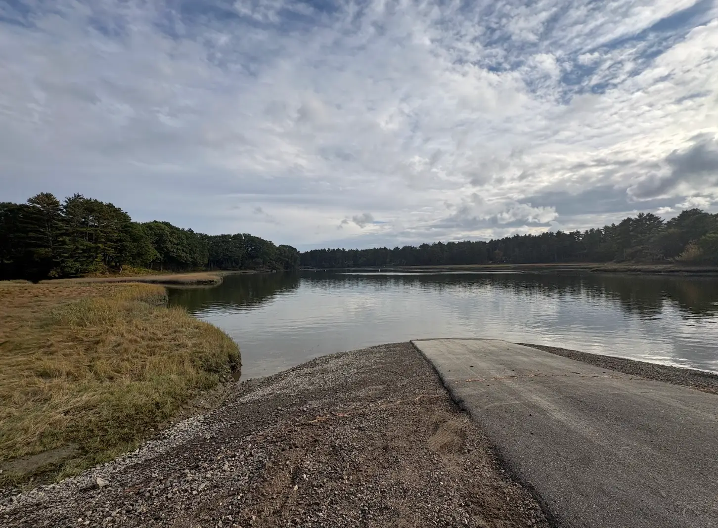Concrete boat ramp leads into a calm forest-lined inlet beneath a cloudy sky.
