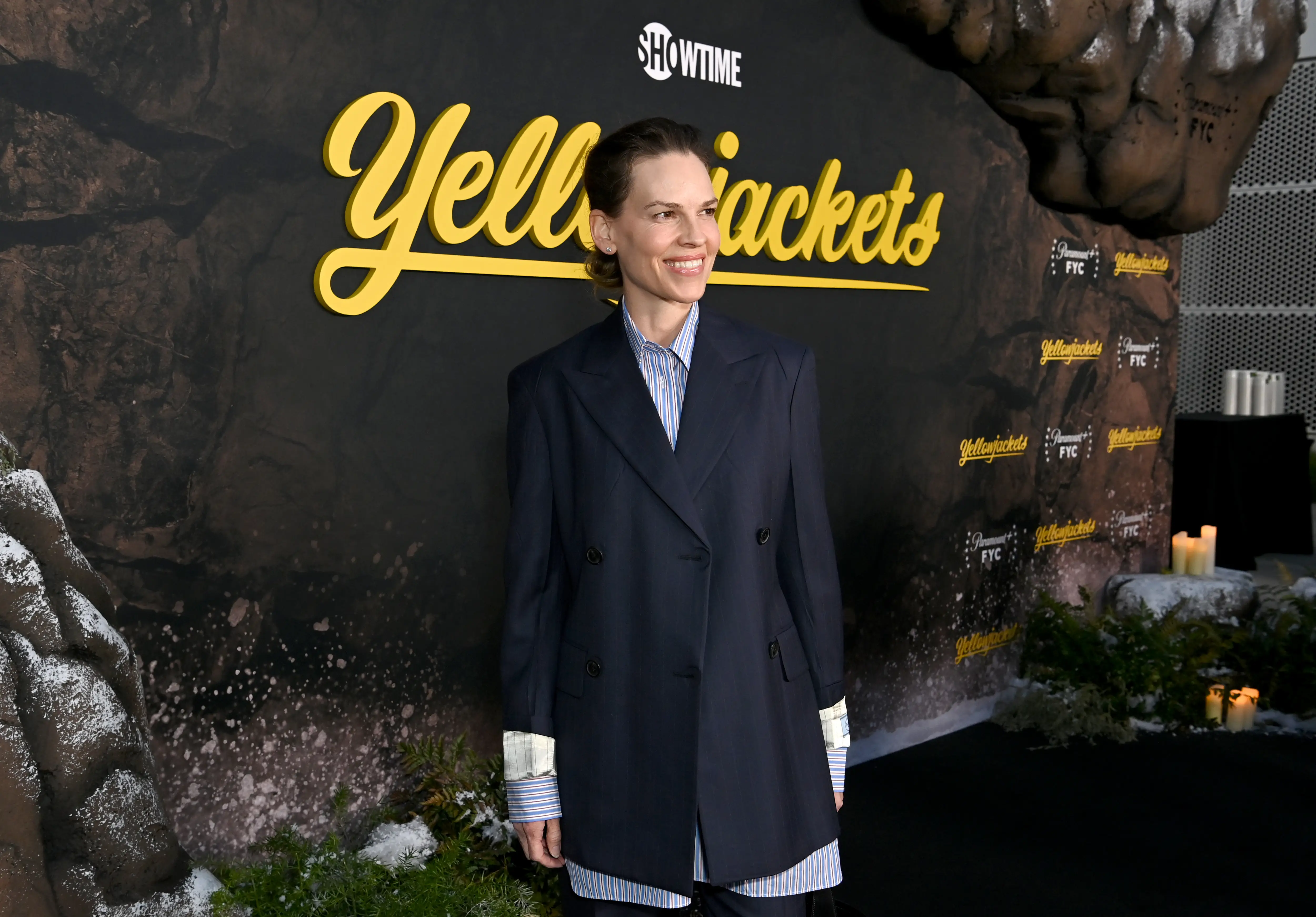 Hilary Swank poses on a dark carpet before a SHOWTIME Yellowjackets backdrop with forest-themed decor.