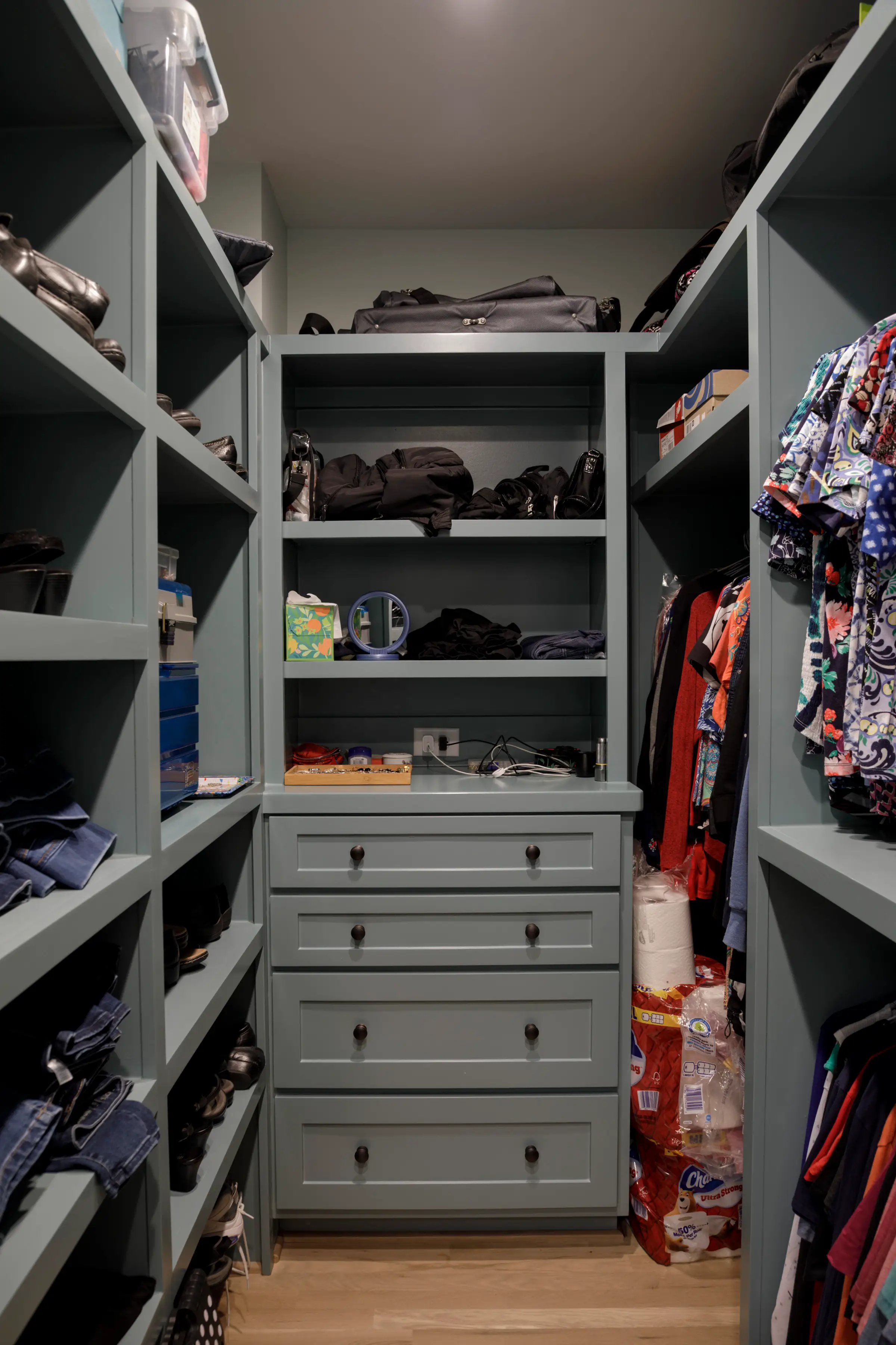 Walk-in closet with gray shelving, drawers, hanging clothes, shoes, bags, and stacked toilet paper packages.
