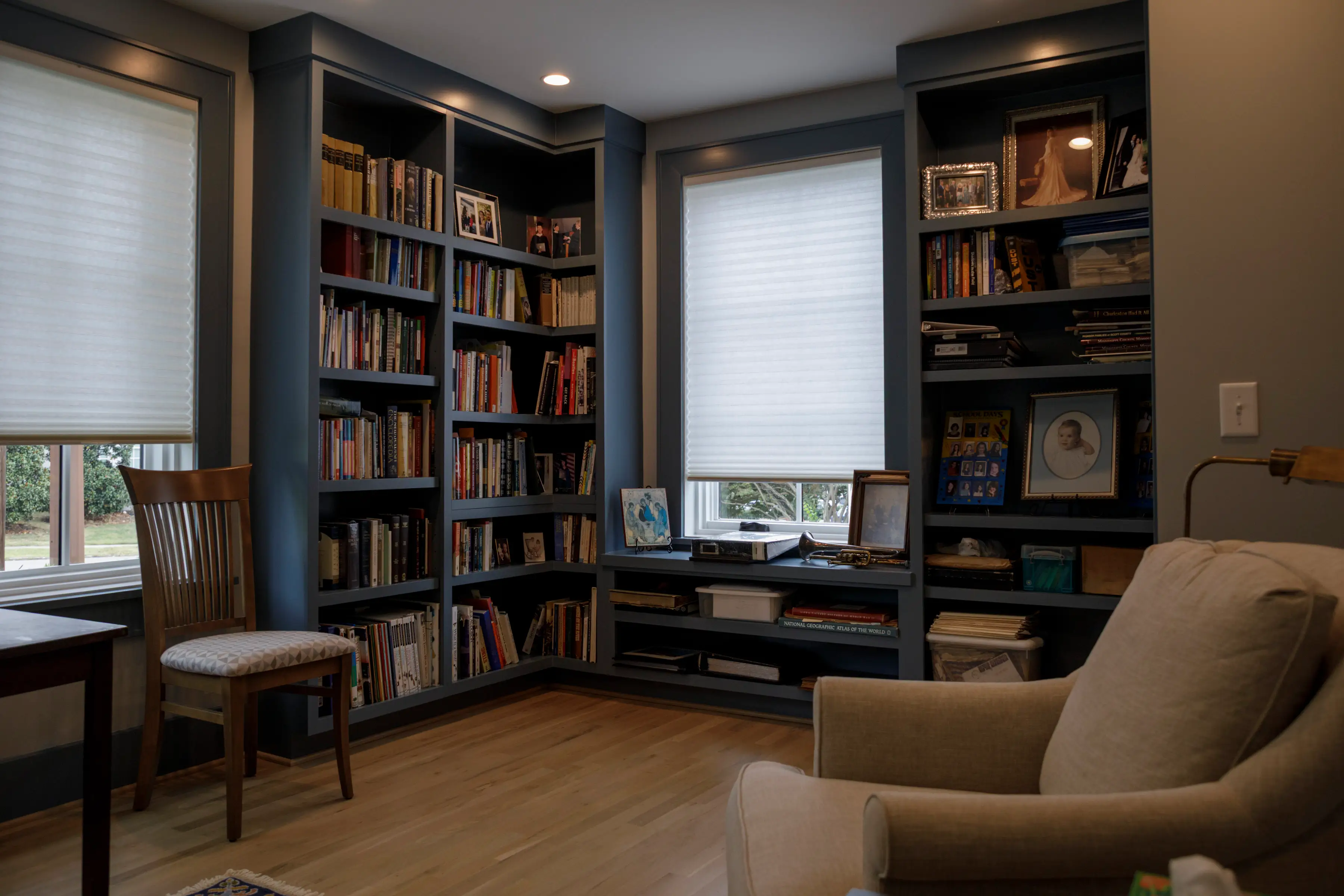 Cozy home library with built-in bookshelves, shaded windows, framed photos, a wooden chair, and a beige armchair.