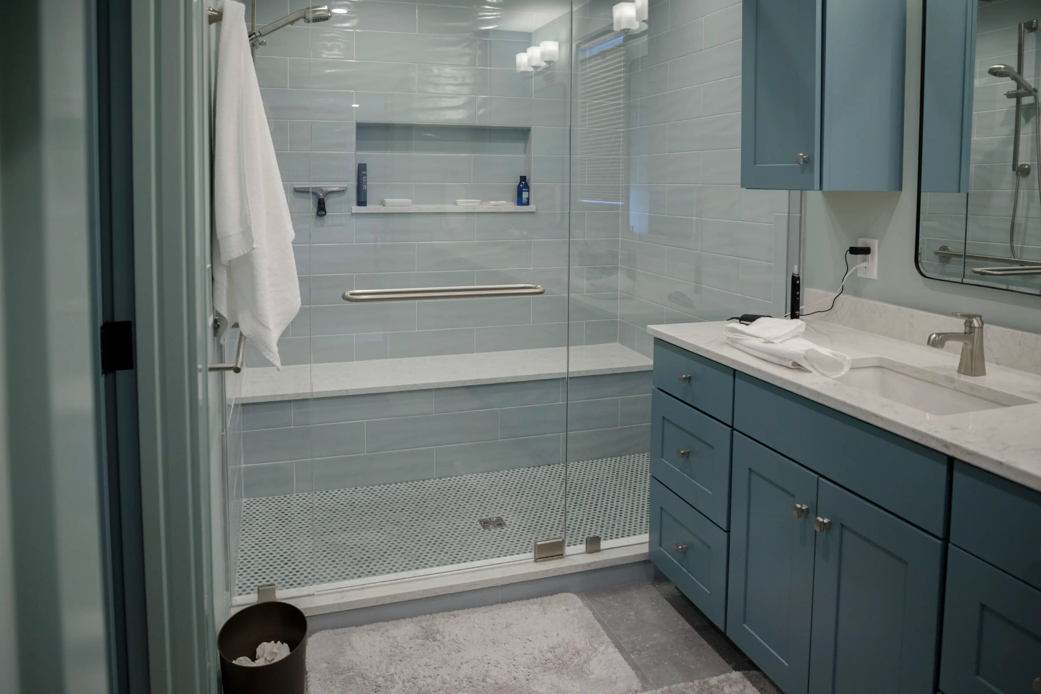 Modern blue bathroom with glass walk-in shower, vanity sink, white towels, and light blue tile walls.