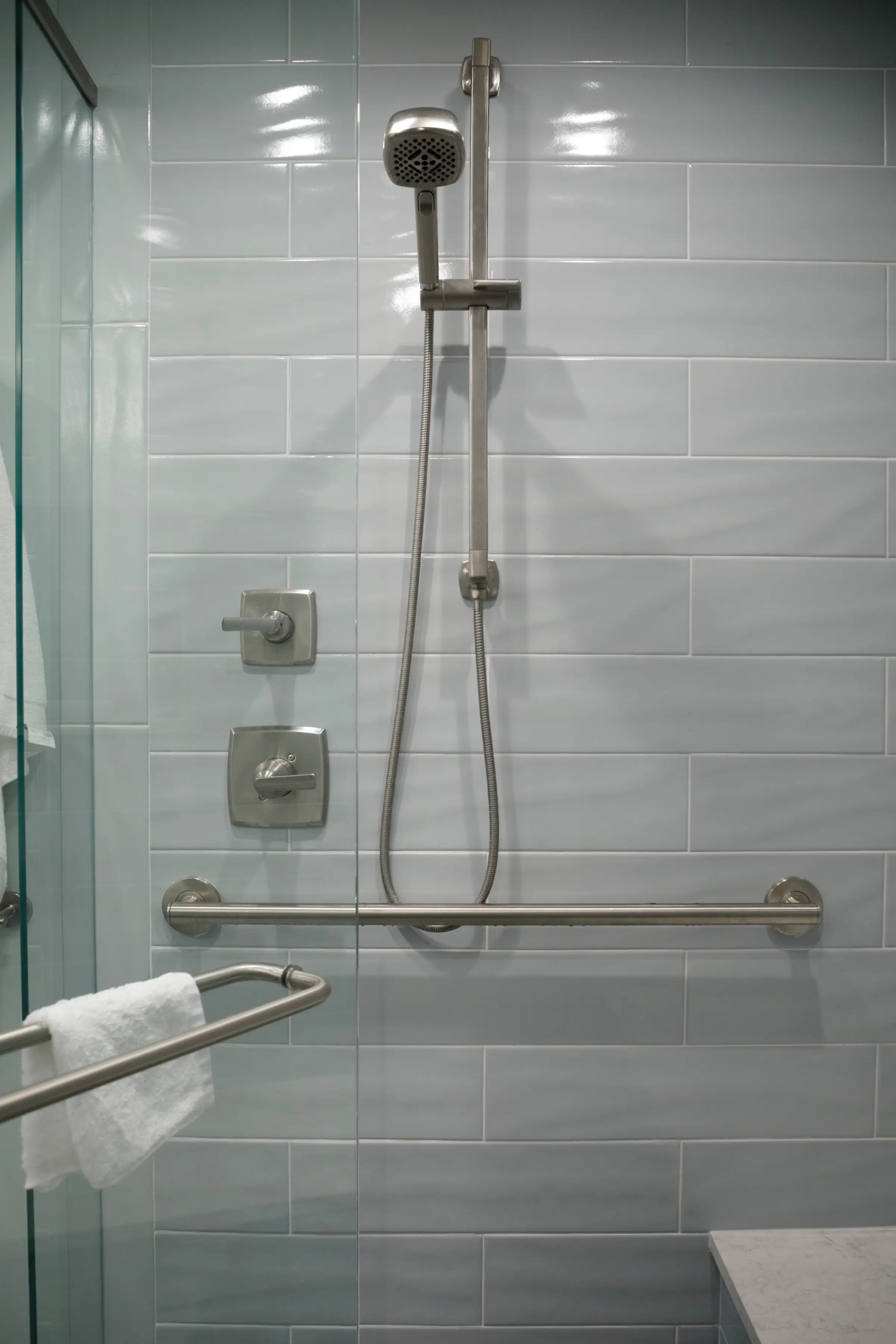 Accessible shower with handheld showerhead, grab bars, pale blue tile walls, glass door, and white towel.