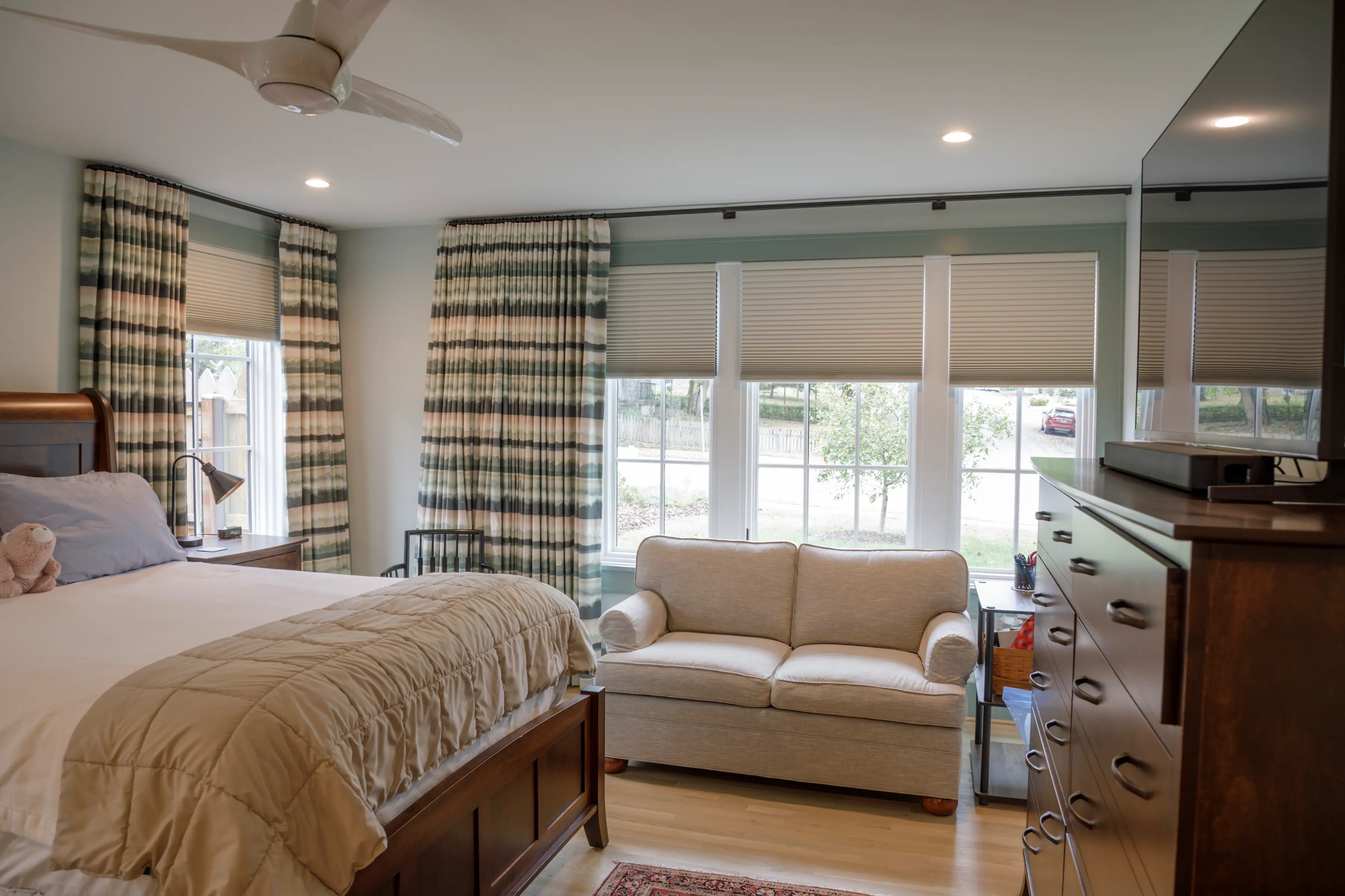 Bright bedroom with a wooden bed, beige loveseat, large curtained windows, dresser, ceiling fan, and recessed lighting.