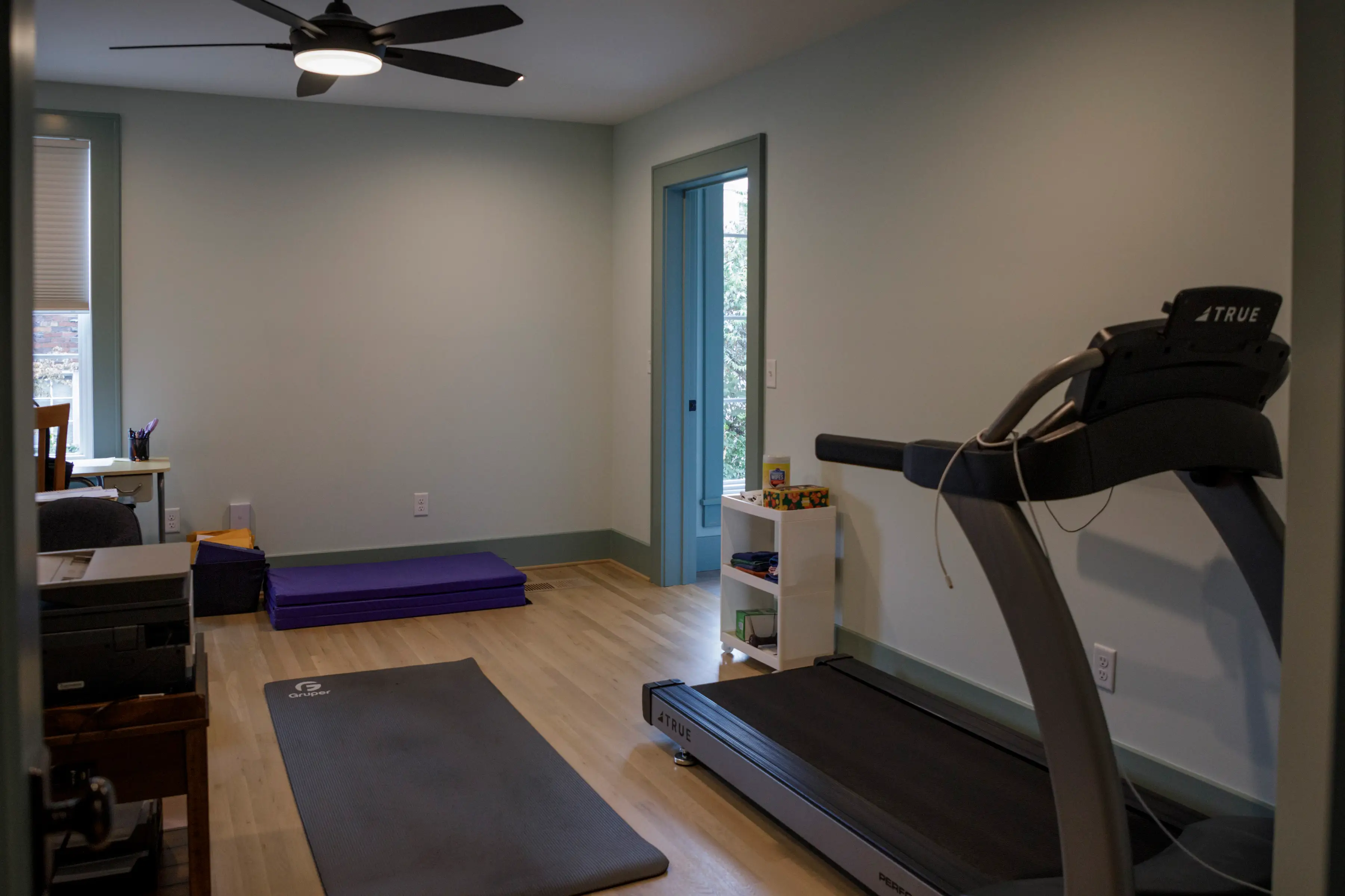 Small home exercise room with a treadmill, floor mats, ceiling fan, desk, and open doorway.