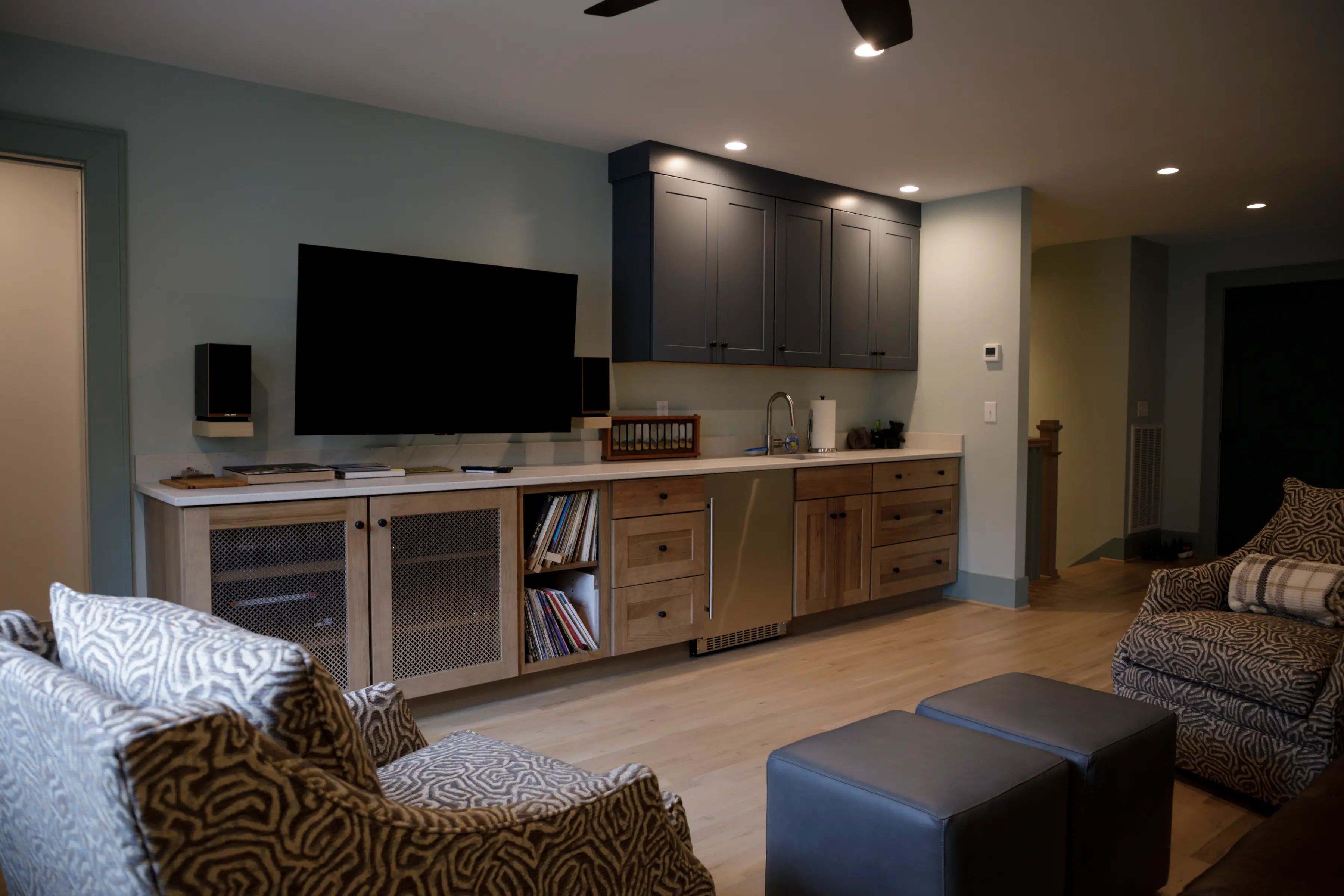 Finished basement lounge with patterned armchairs, wall-mounted TV, built-in cabinets, sink, and under-counter beverage cooler.
