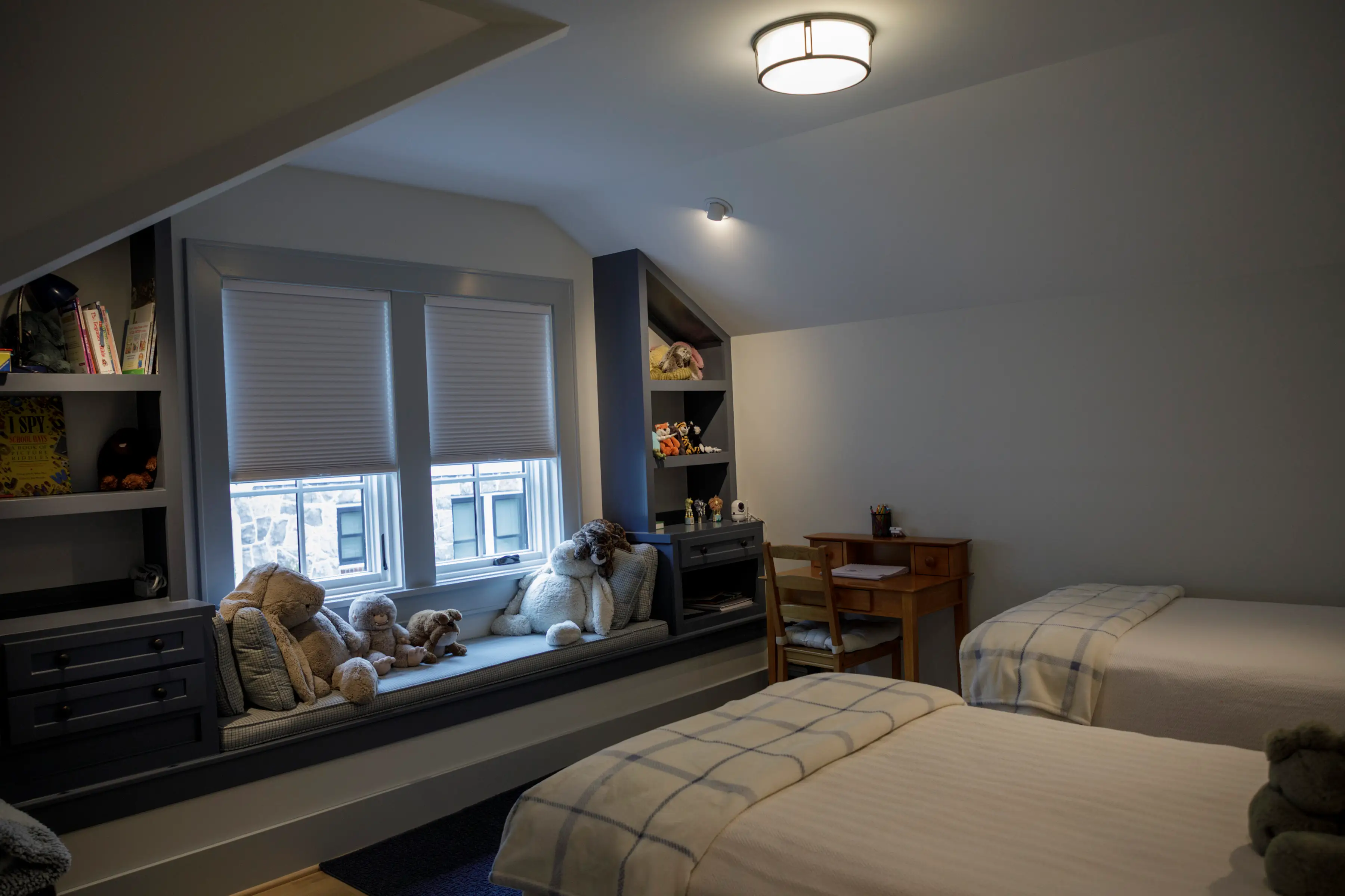 Attic-style bedroom with twin beds, built-in window seat, stuffed animals, shelves, desk, and closed blinds.