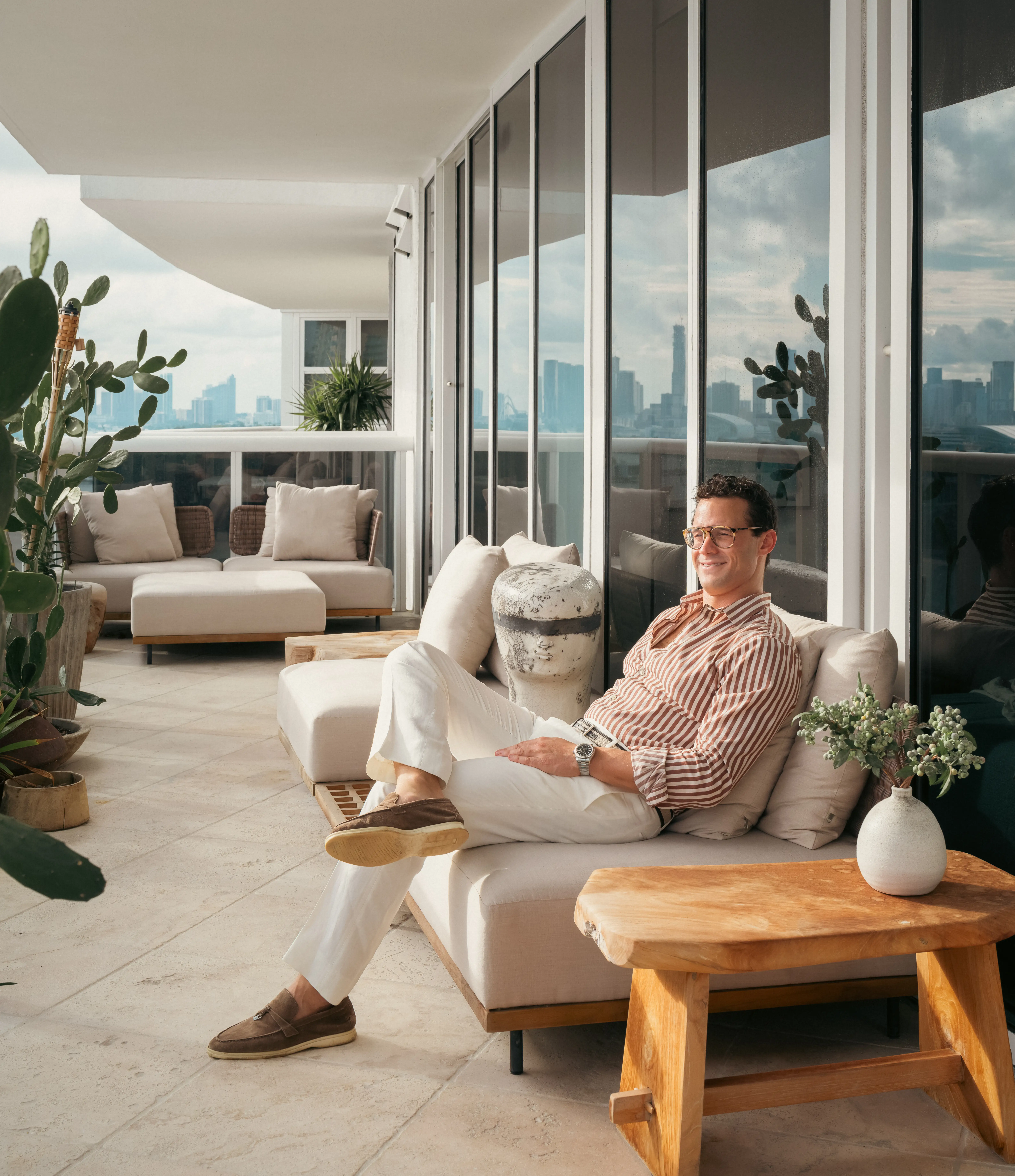 Person in glasses relaxes on a cushioned balcony sofa overlooking a distant city skyline.