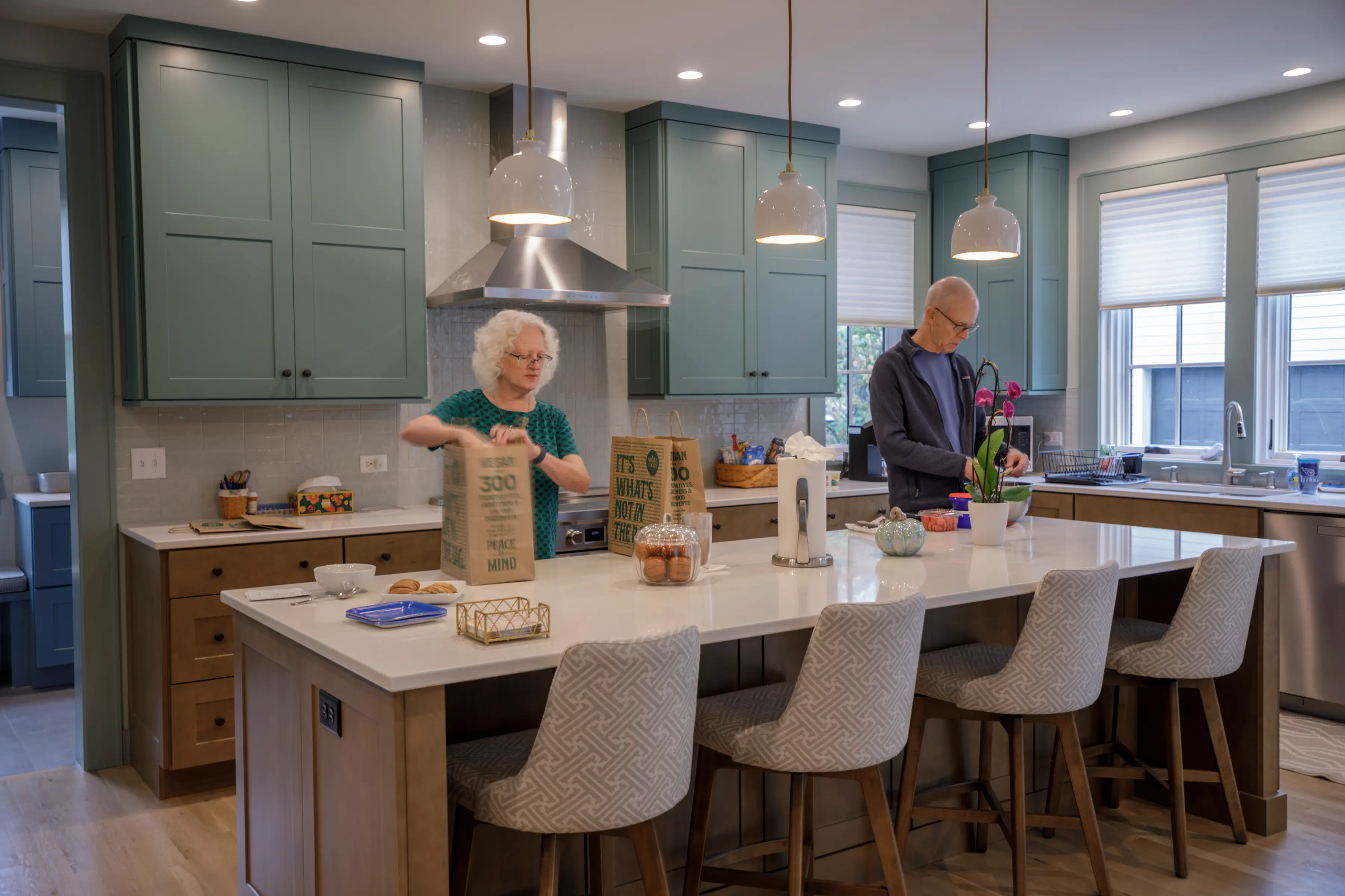 Two people prepare paper bags on a kitchen island in a bright modern kitchen with green cabinets.