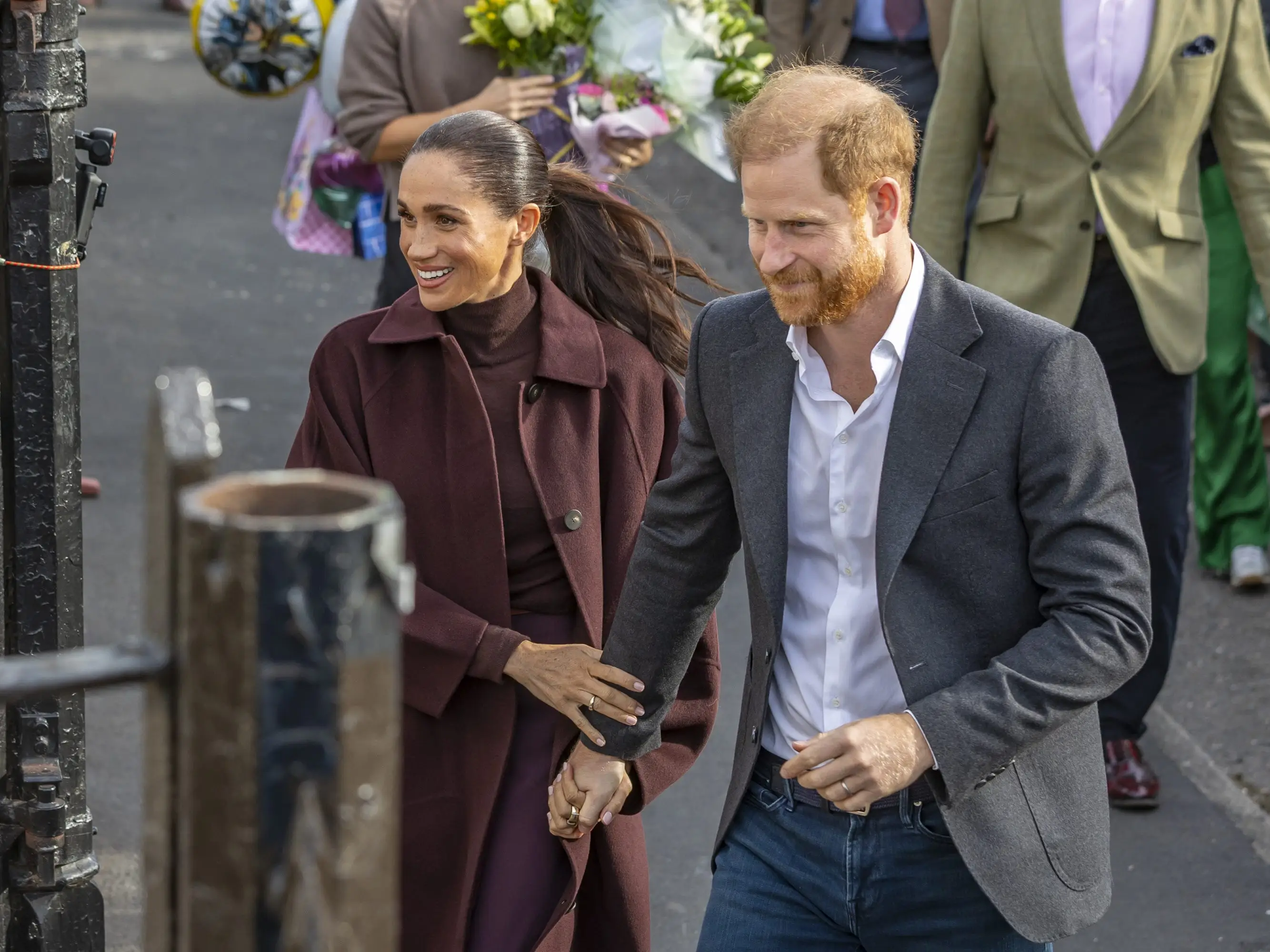 Meghan Markle and Prince Harry walk hand in hand on a city street with people behind them.