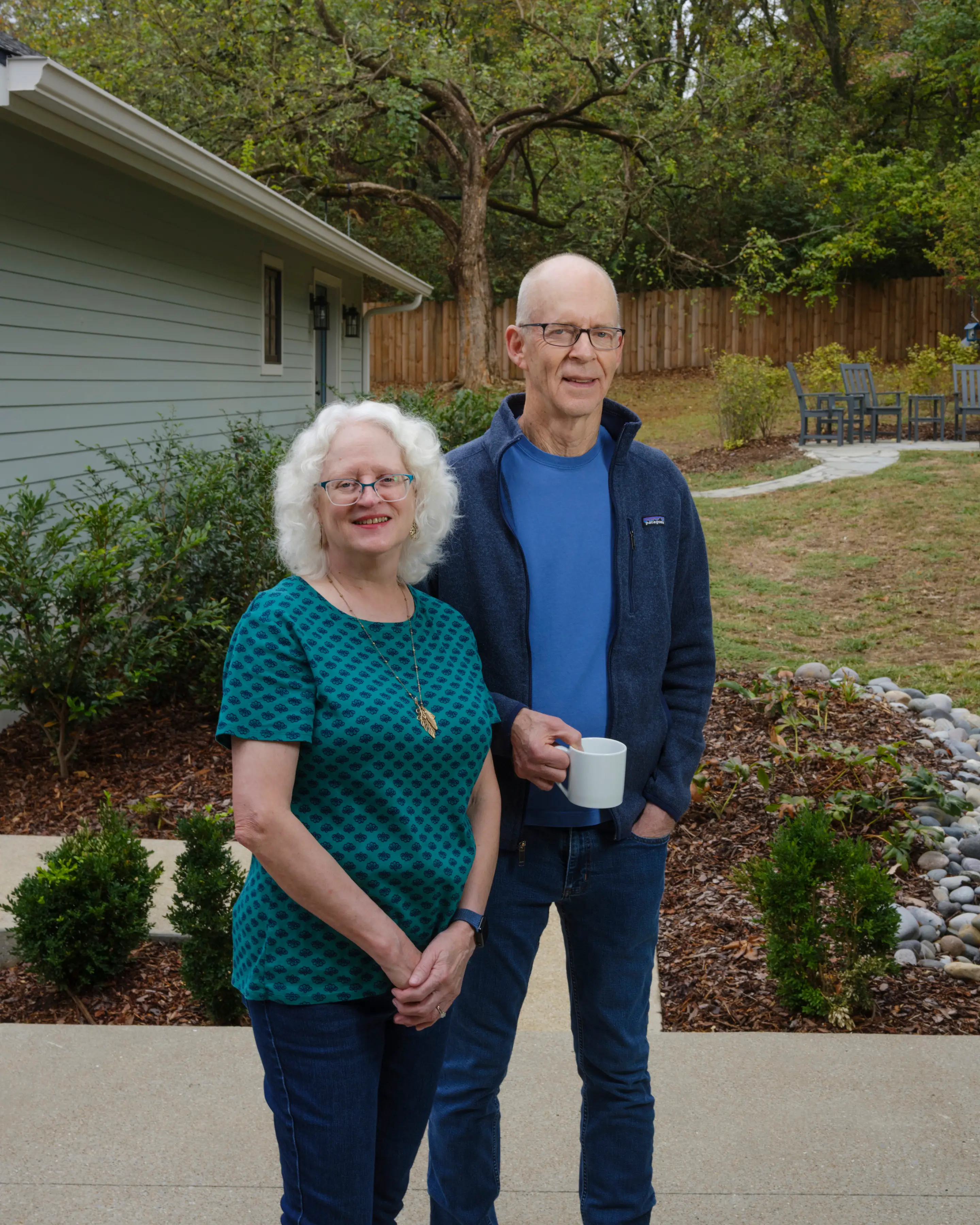 Two people stand in a landscaped backyard beside a house, with one holding a white mug.