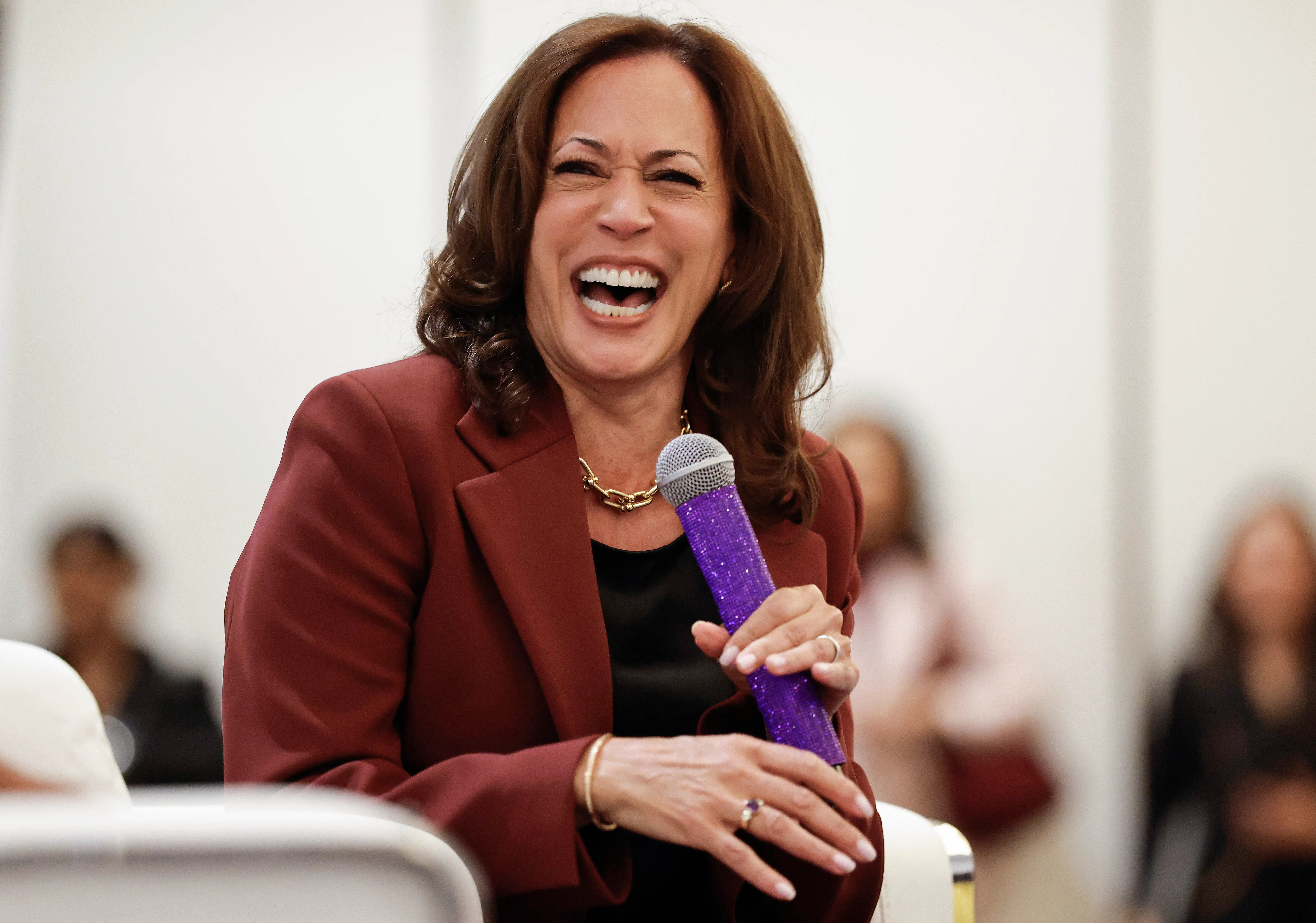 Kamala Harris laughs while holding a purple microphone during an indoor seated event.