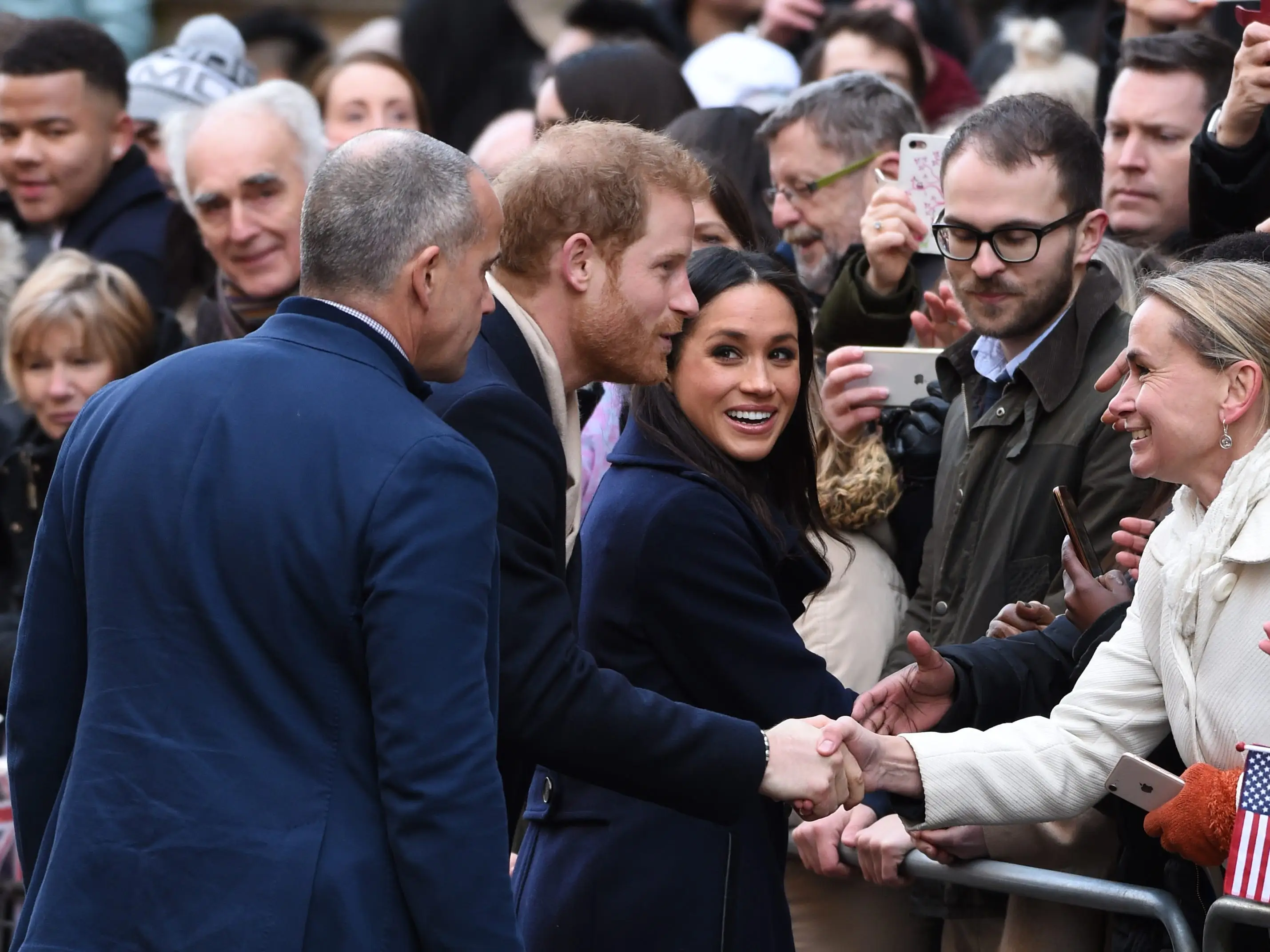 Prince Harry and Meghan Markle greet and shake hands with a crowd behind a metal barricade.