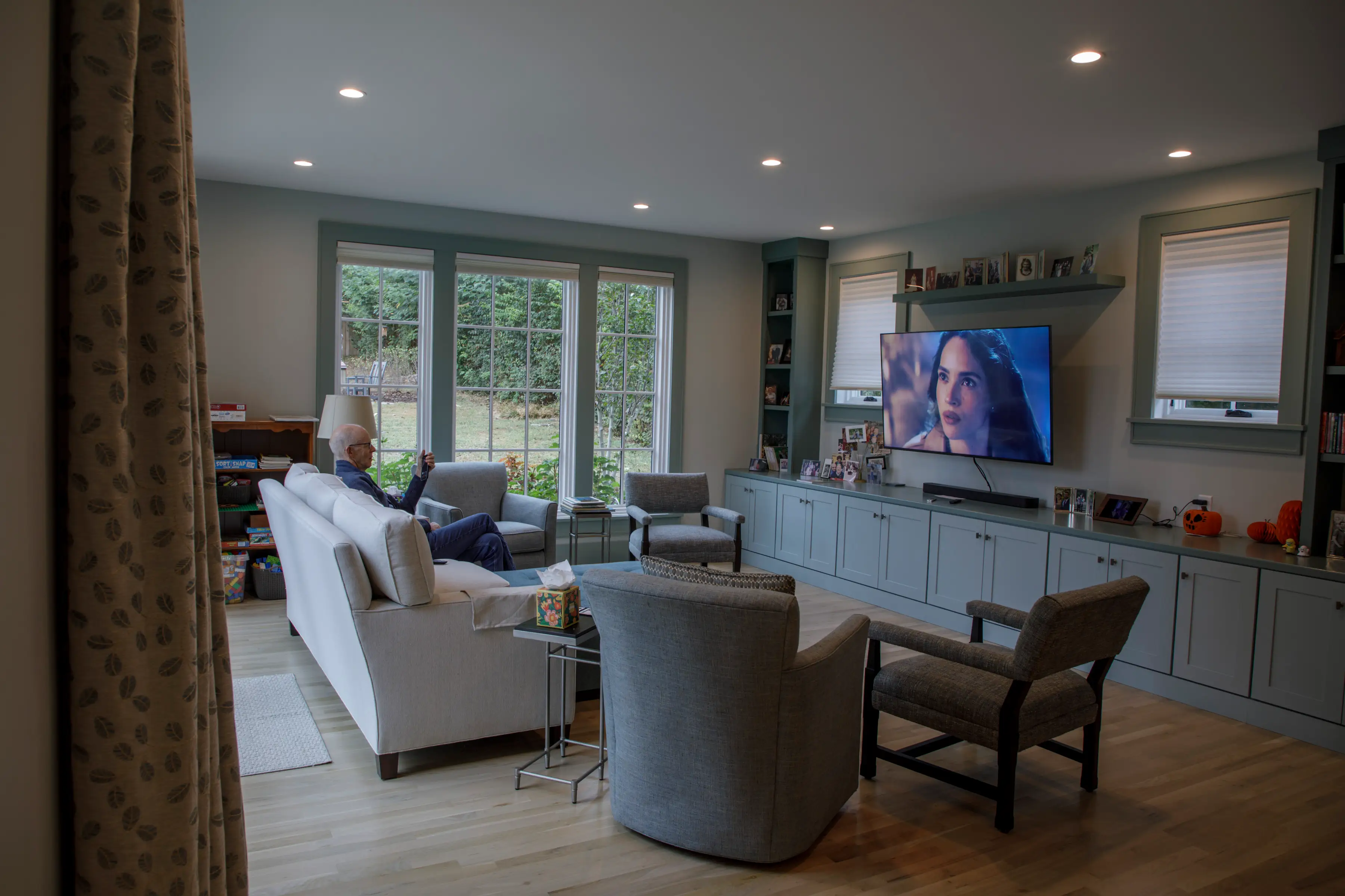 A person sits on a sofa watching a wall-mounted television in a spacious living room with large windows.