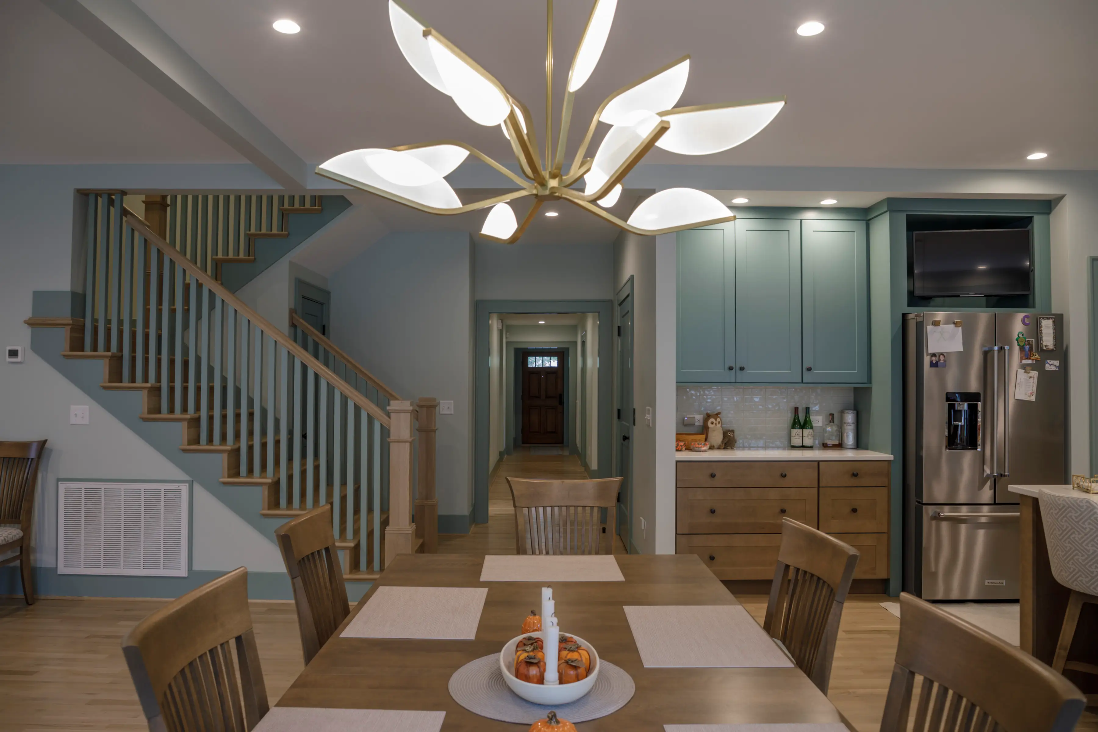 Dining table beneath a modern chandelier beside a staircase and teal kitchen cabinetry in an open-plan home.
