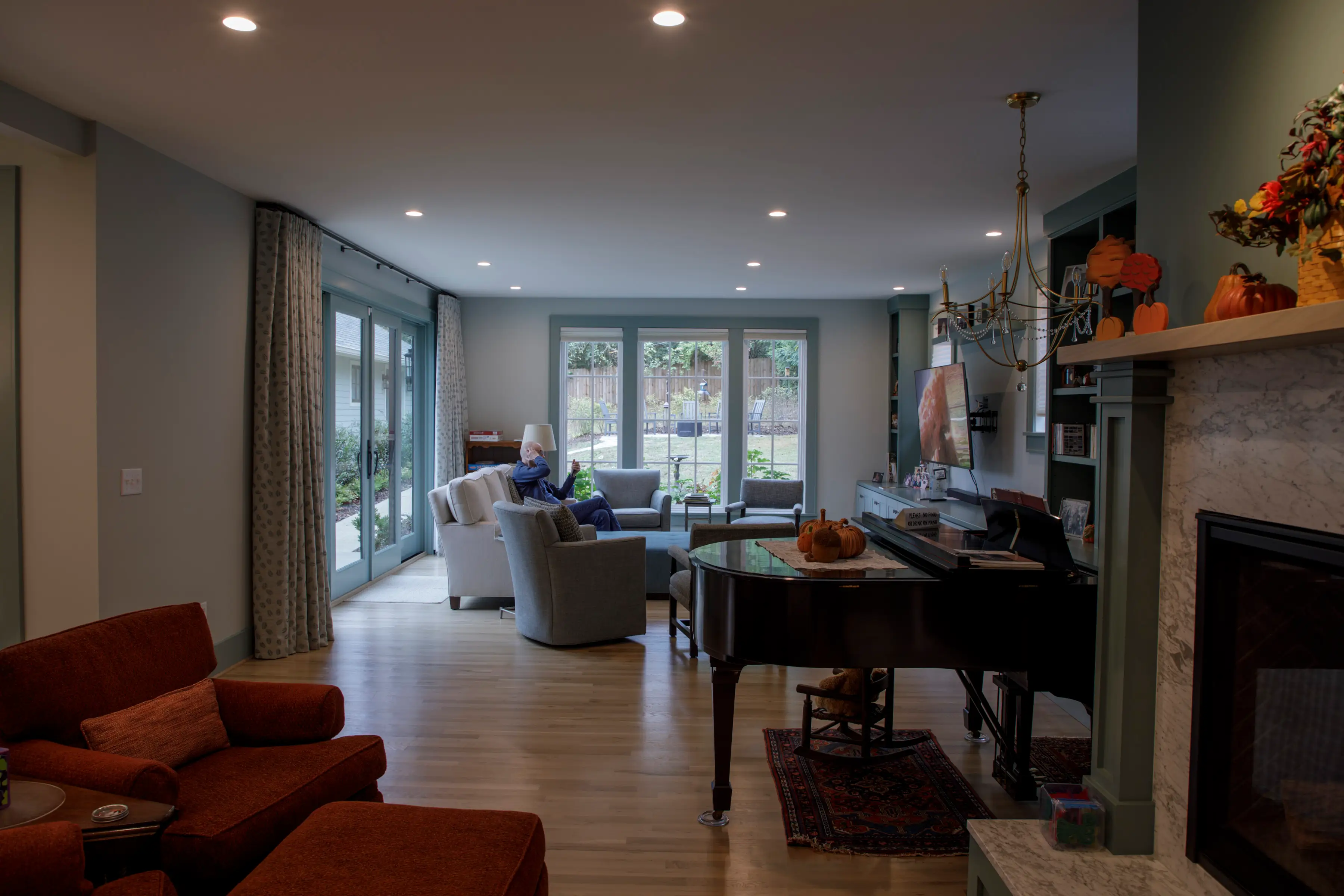 A person sits on a sofa in a spacious living room with a grand piano, fireplace, and autumn decorations.