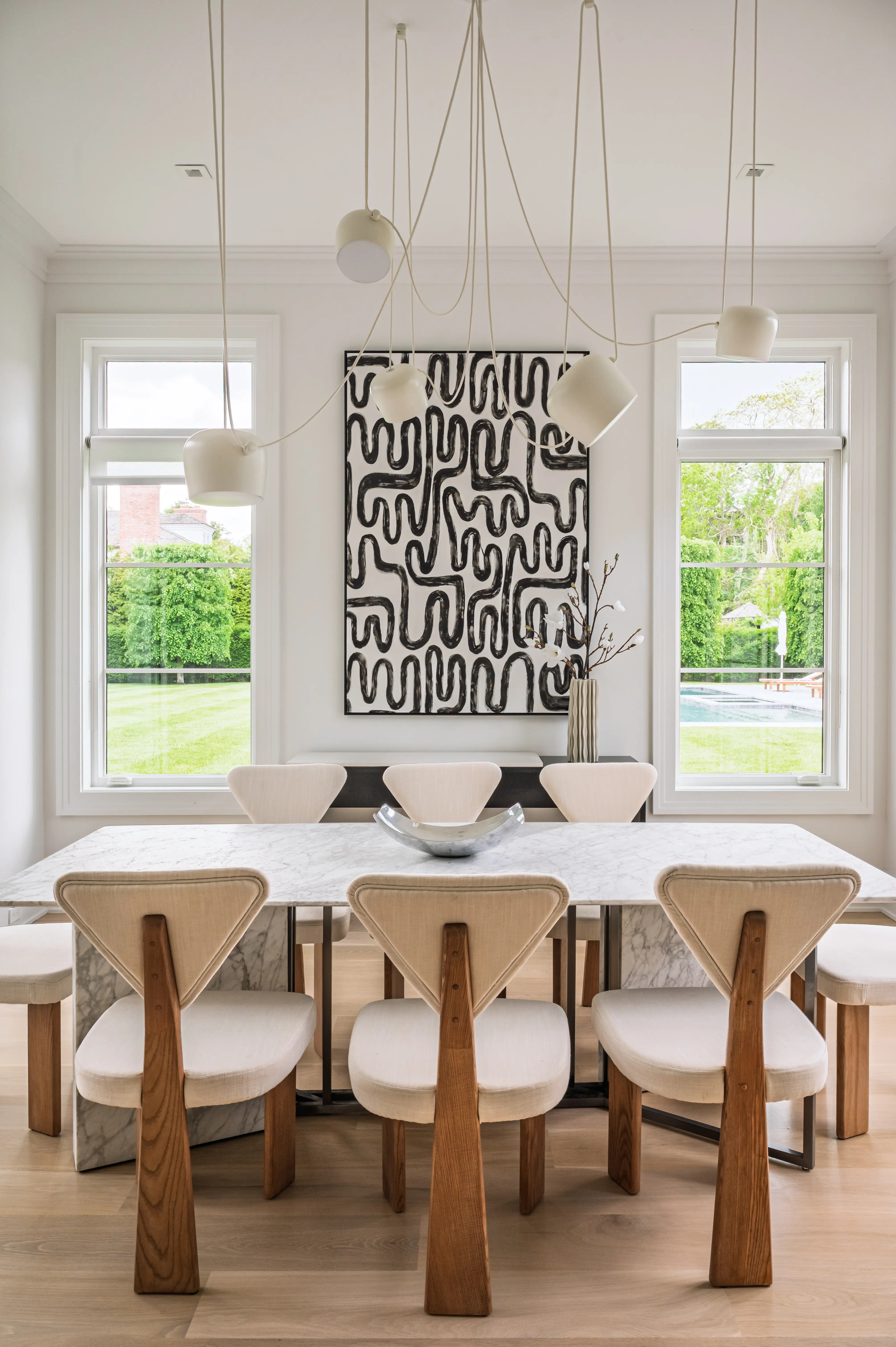 Bright dining room with marble table, upholstered wooden chairs, hanging pendant lights, and abstract black-and-white wall art.