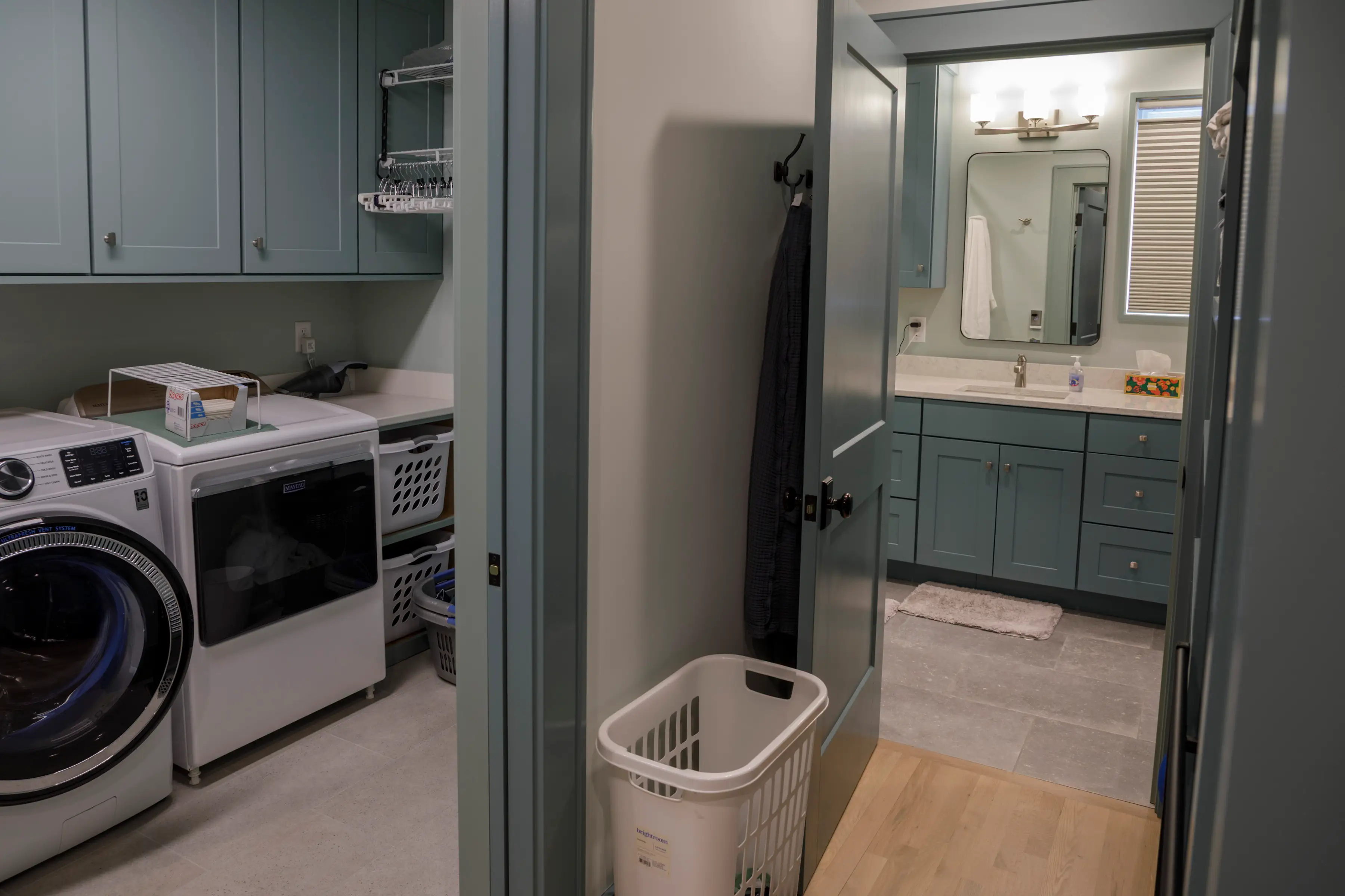 Laundry room with washer, dryer and baskets sits beside a bathroom with blue-green cabinets and vanity.