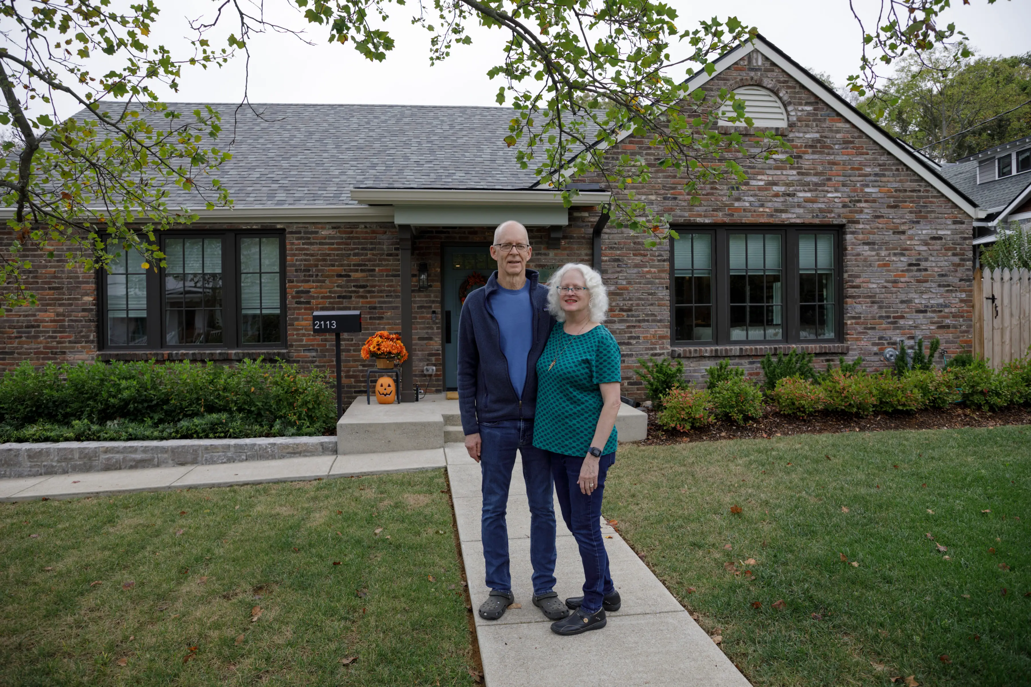 Two people stand on a front walkway outside a brick house with pumpkins and the address number 2113 visible.