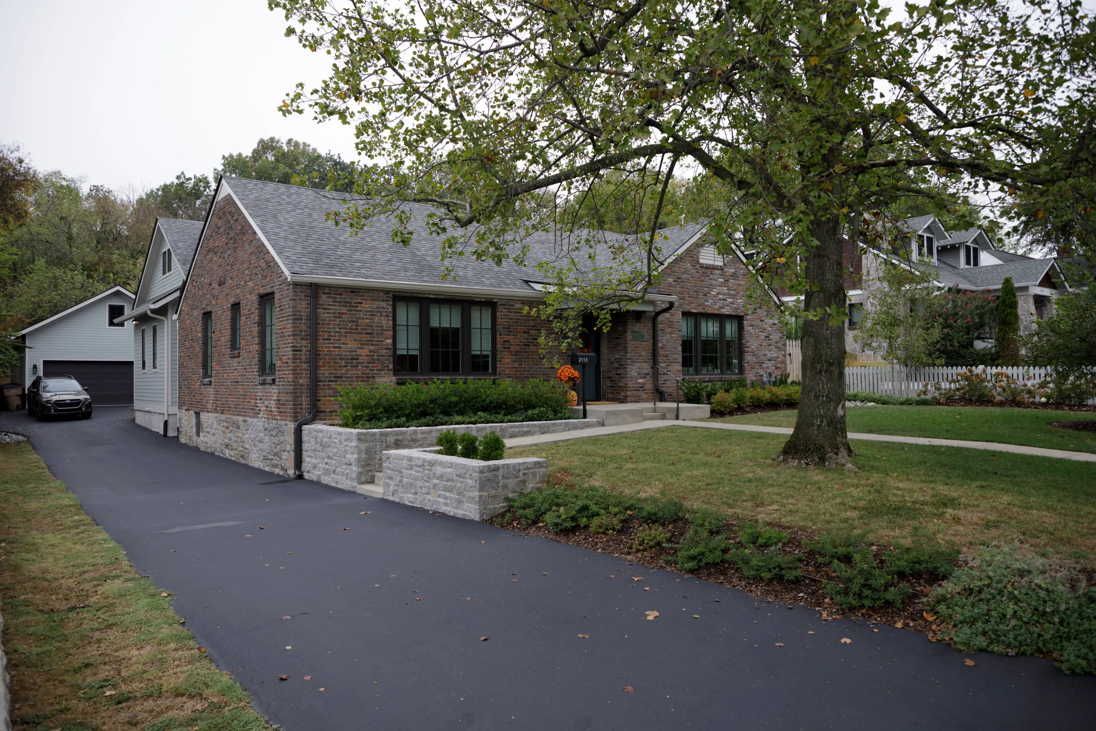 Brick suburban house with a paved driveway, front lawn, mature tree, and landscaped garden beds.
