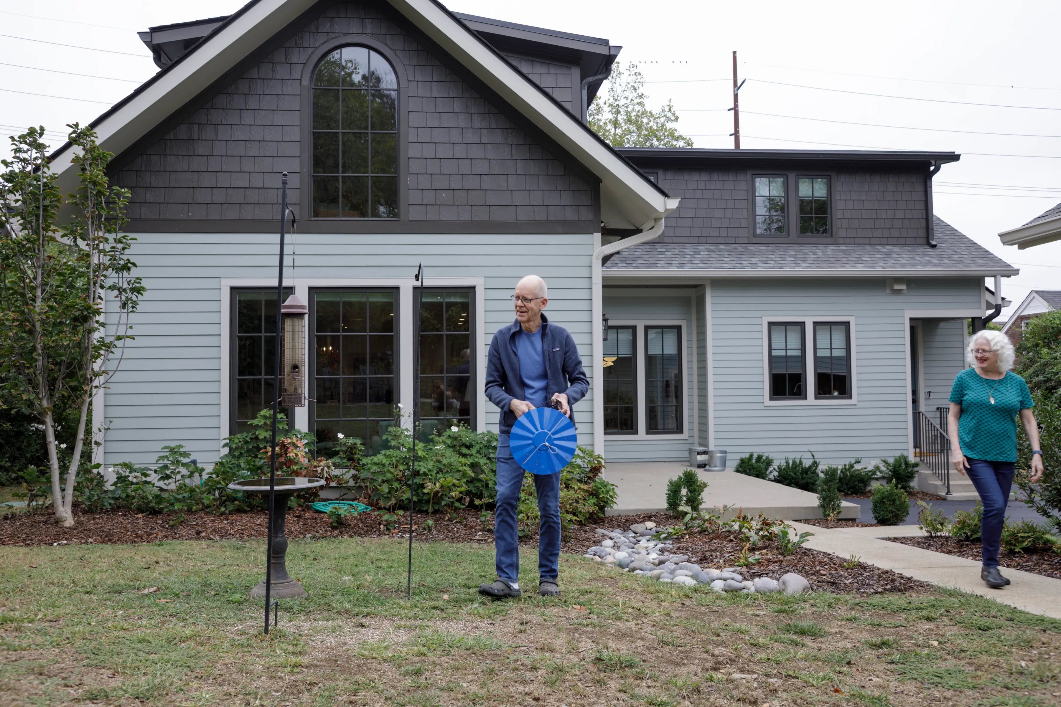 Two people stand in the landscaped yard of a gray two-story house with large windows and a bird feeder.
