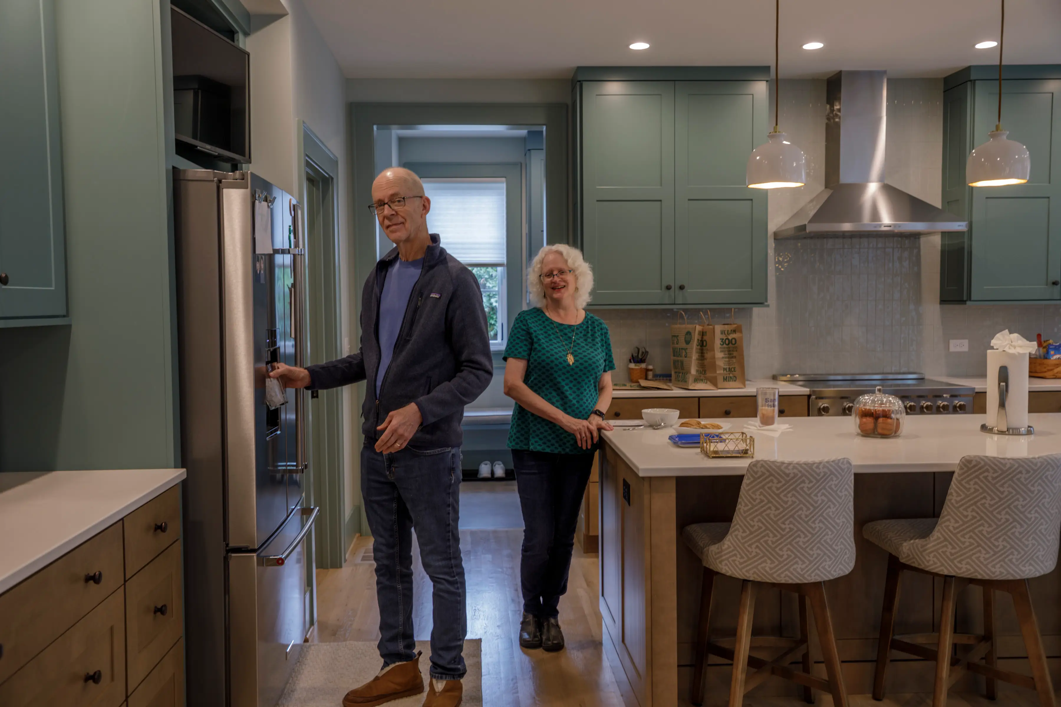 Two people stand in a renovated kitchen with green cabinets, stainless appliances, island seating, and pendant lights.