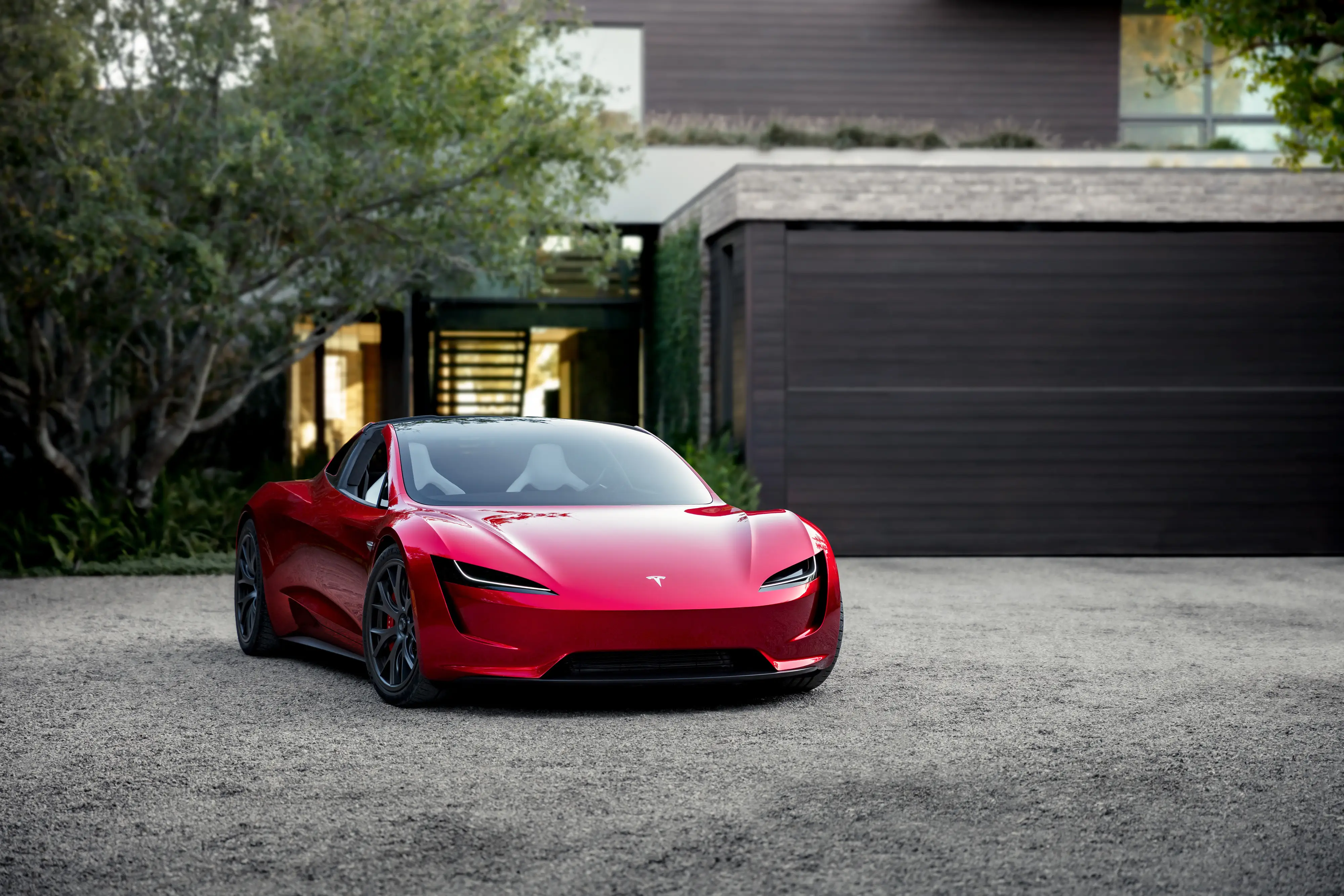 A Tesla Roadster prototype is parked on a gravel driveway in front of a modern contemporary home.