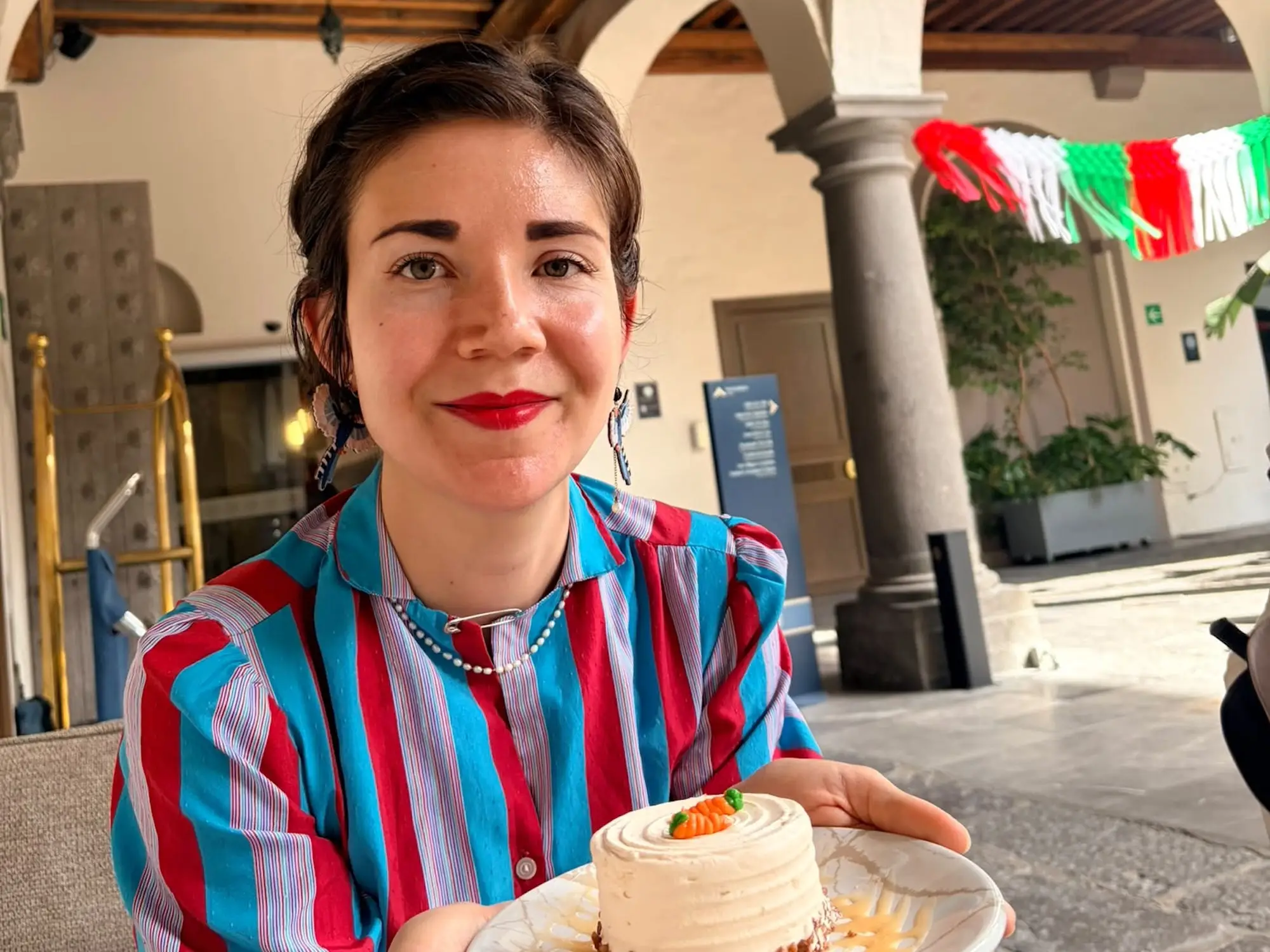 Person in a striped shirt holds a small frosted dessert in an arched courtyard with colorful bunting.