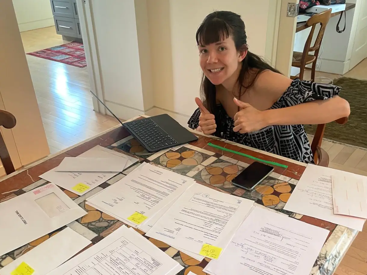 Person gives two thumbs up at a dining table covered with paperwork, sticky notes, a tablet, and a smartphone.
