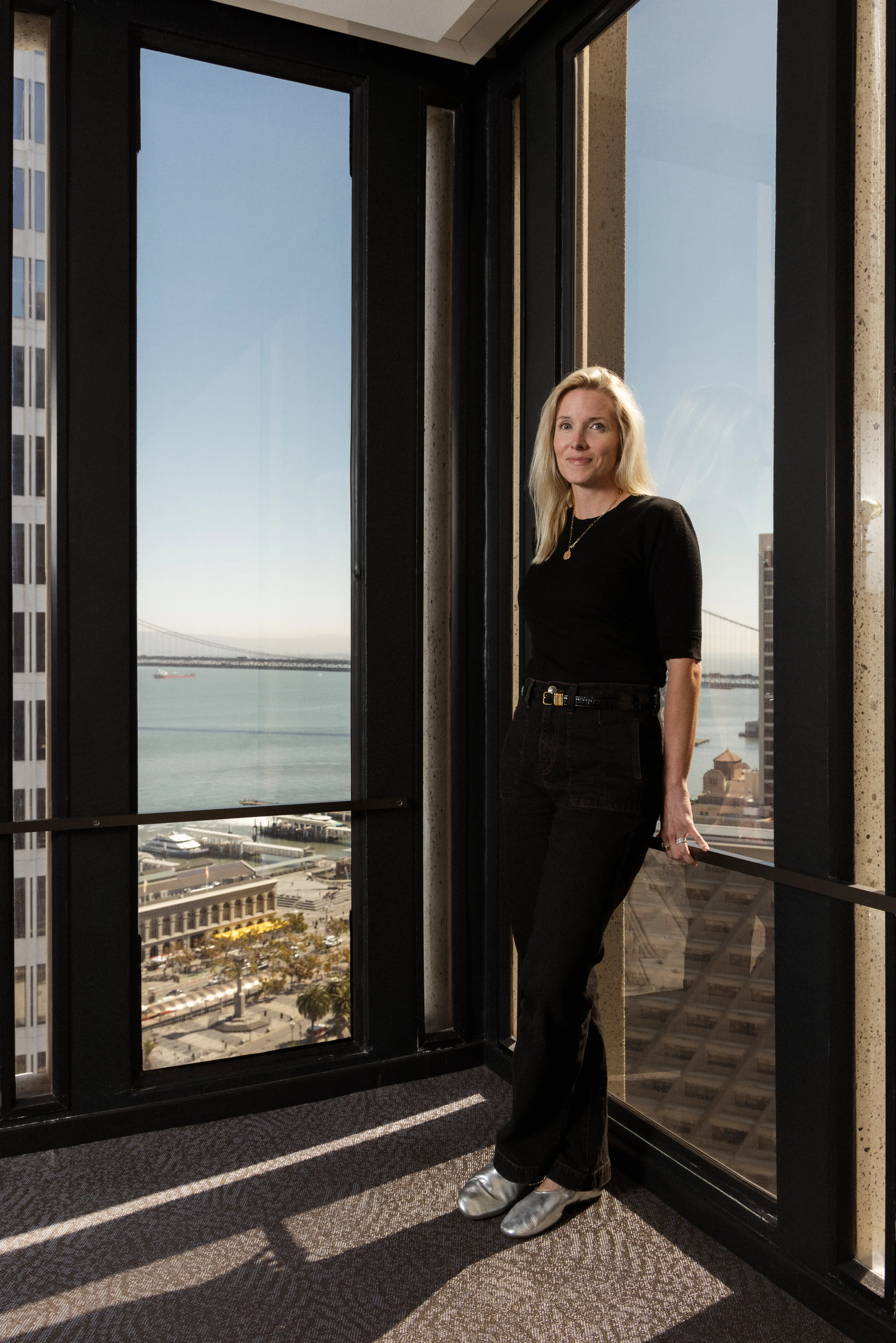 Person stands by floor-to-ceiling office windows overlooking a bay, waterfront, and distant bridge.