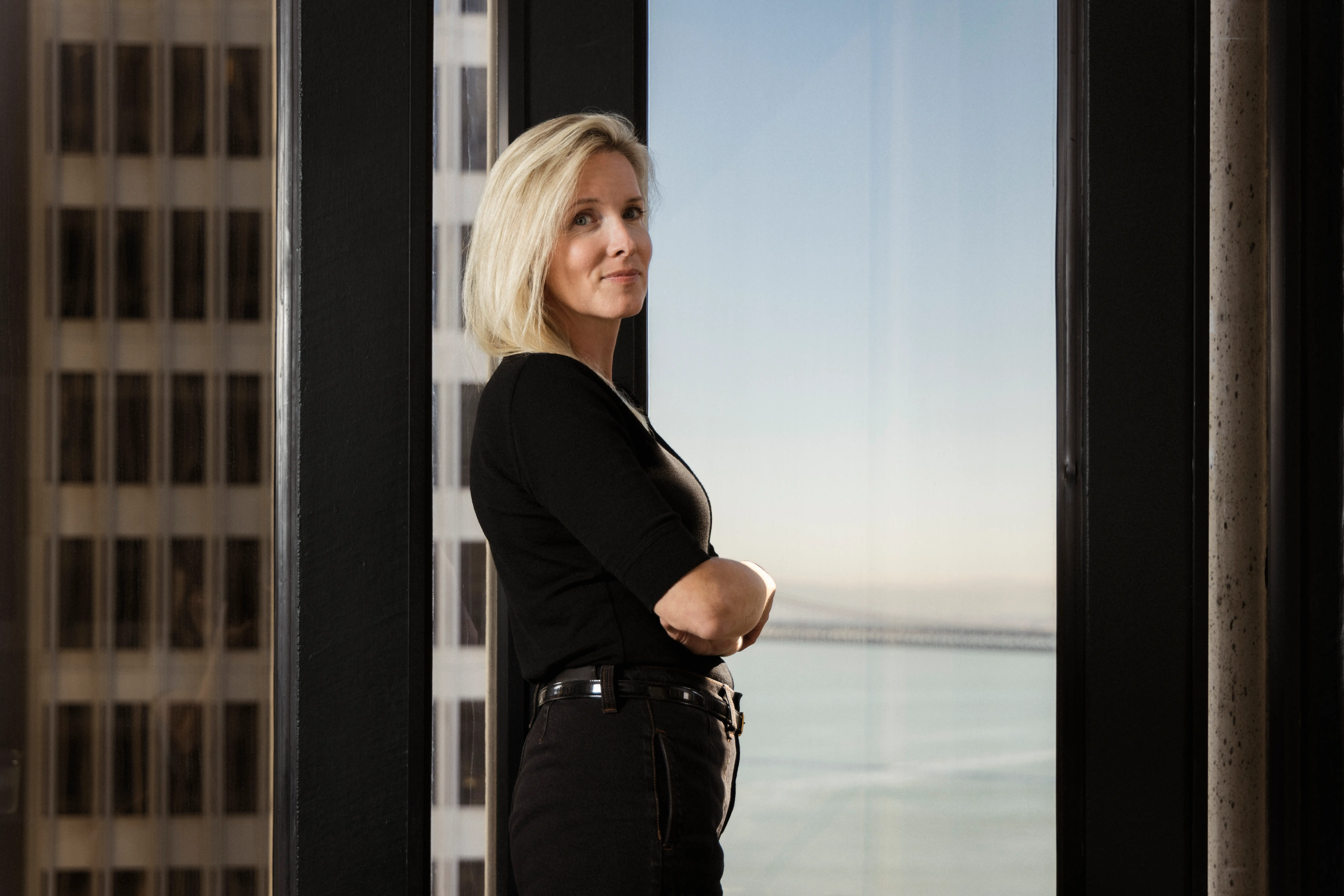Person in black clothing stands with arms crossed beside tall windows overlooking city buildings and water.