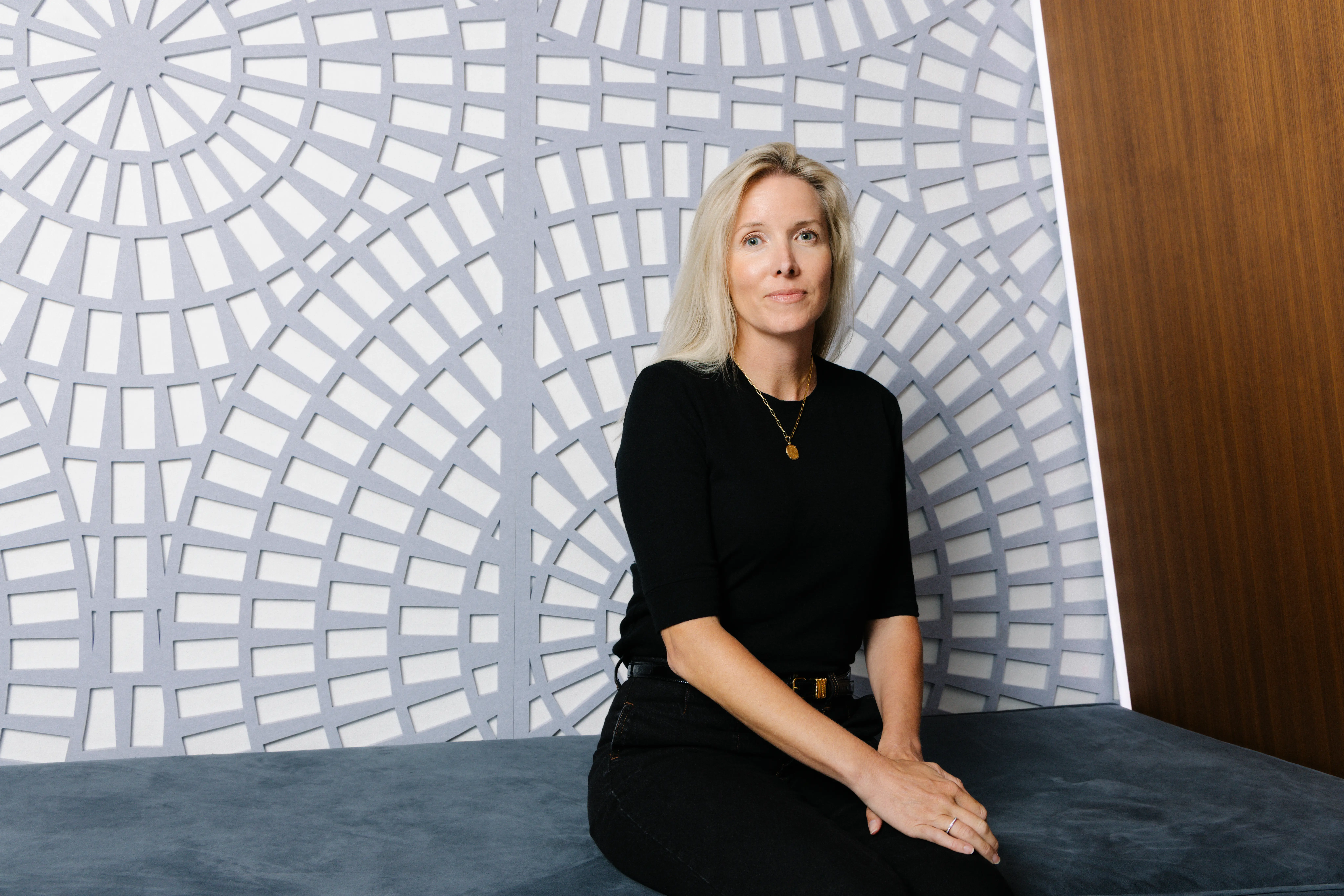 A seated person in black clothing poses beside a patterned wall and wood paneling.