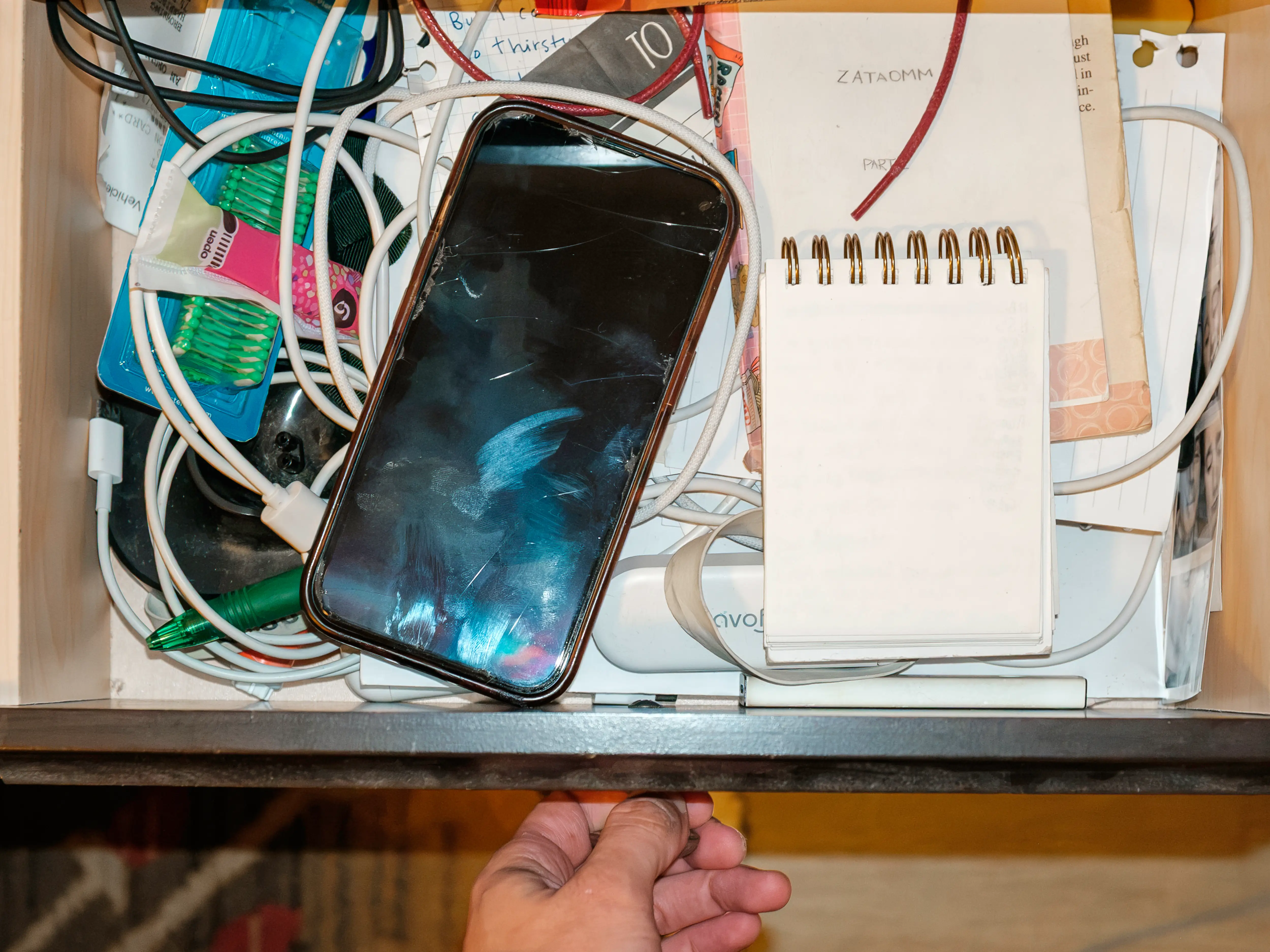 Open drawer holds a scratched smartphone, tangled charging cables, a small spiral notebook, and miscellaneous personal items.