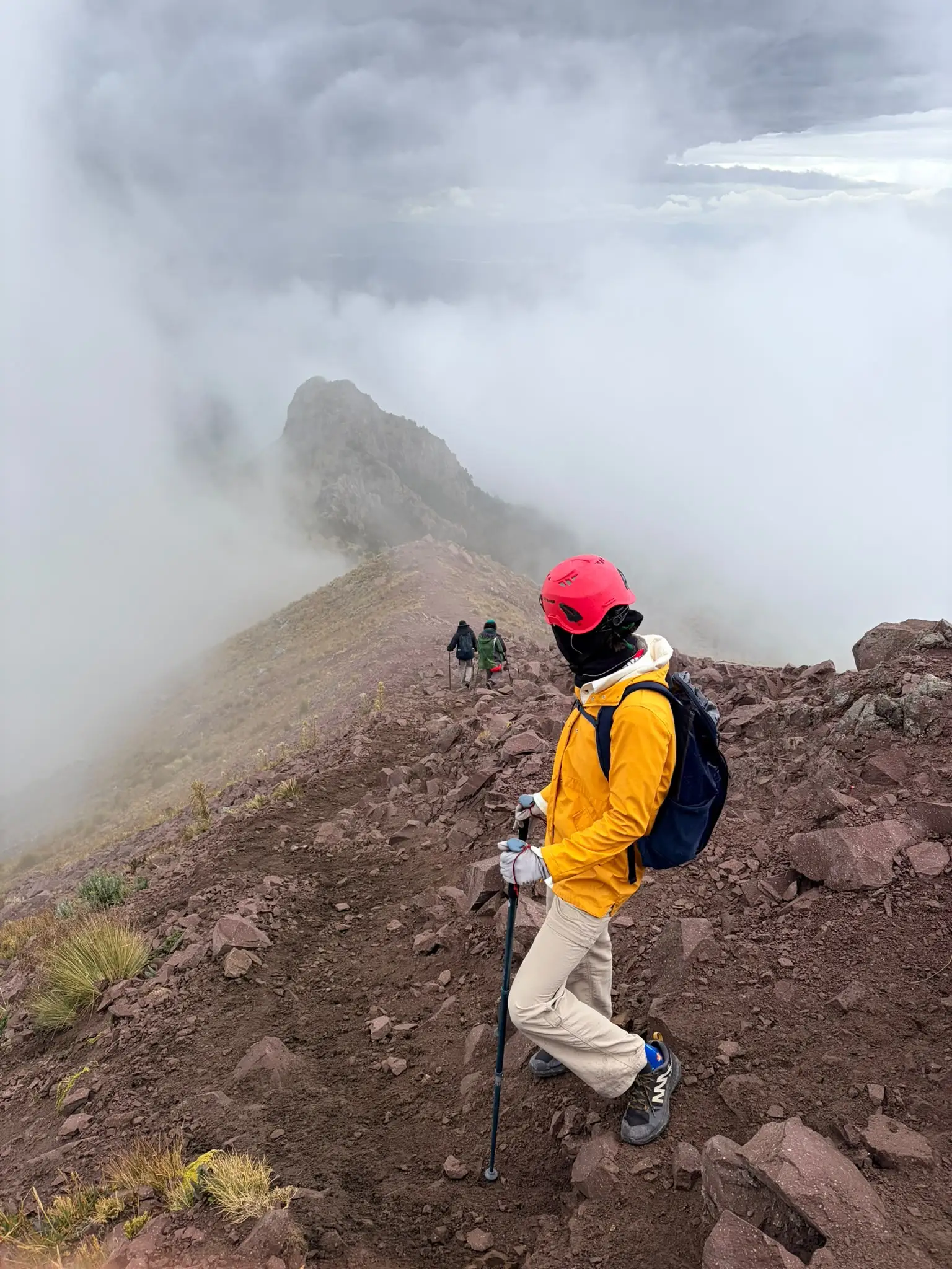 Hikers descend a rocky mountain ridge surrounded by dense clouds and mist, led by a person in a red helmet.