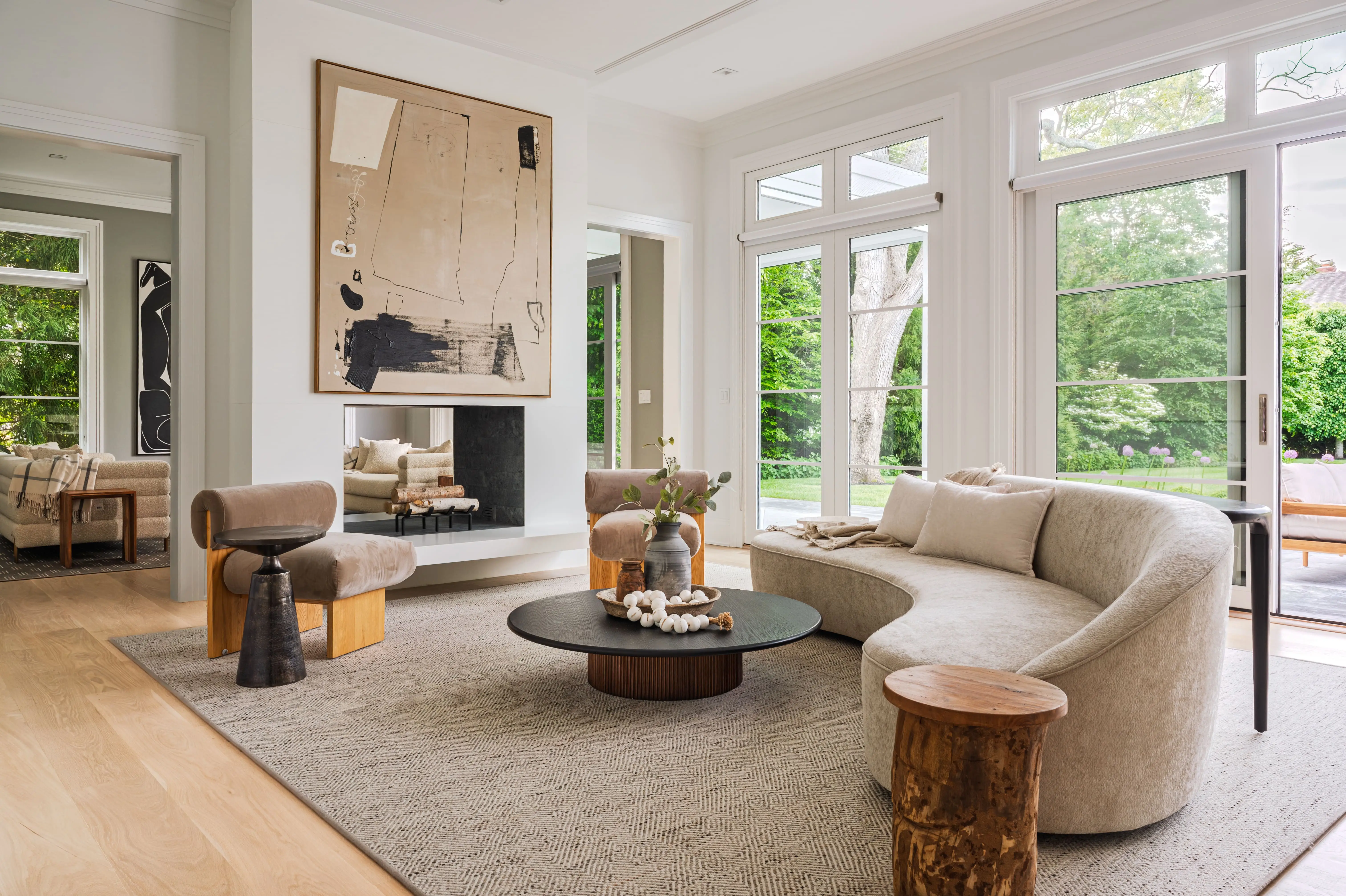 Bright contemporary living room with curved sofa, round coffee table, abstract wall art, fireplace, and large garden-facing windows.