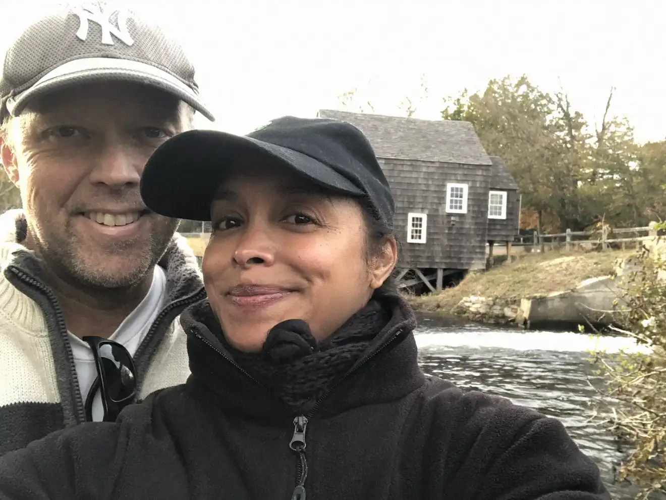 Two people pose for a selfie beside water with a rustic wooden building and autumn trees in the background.