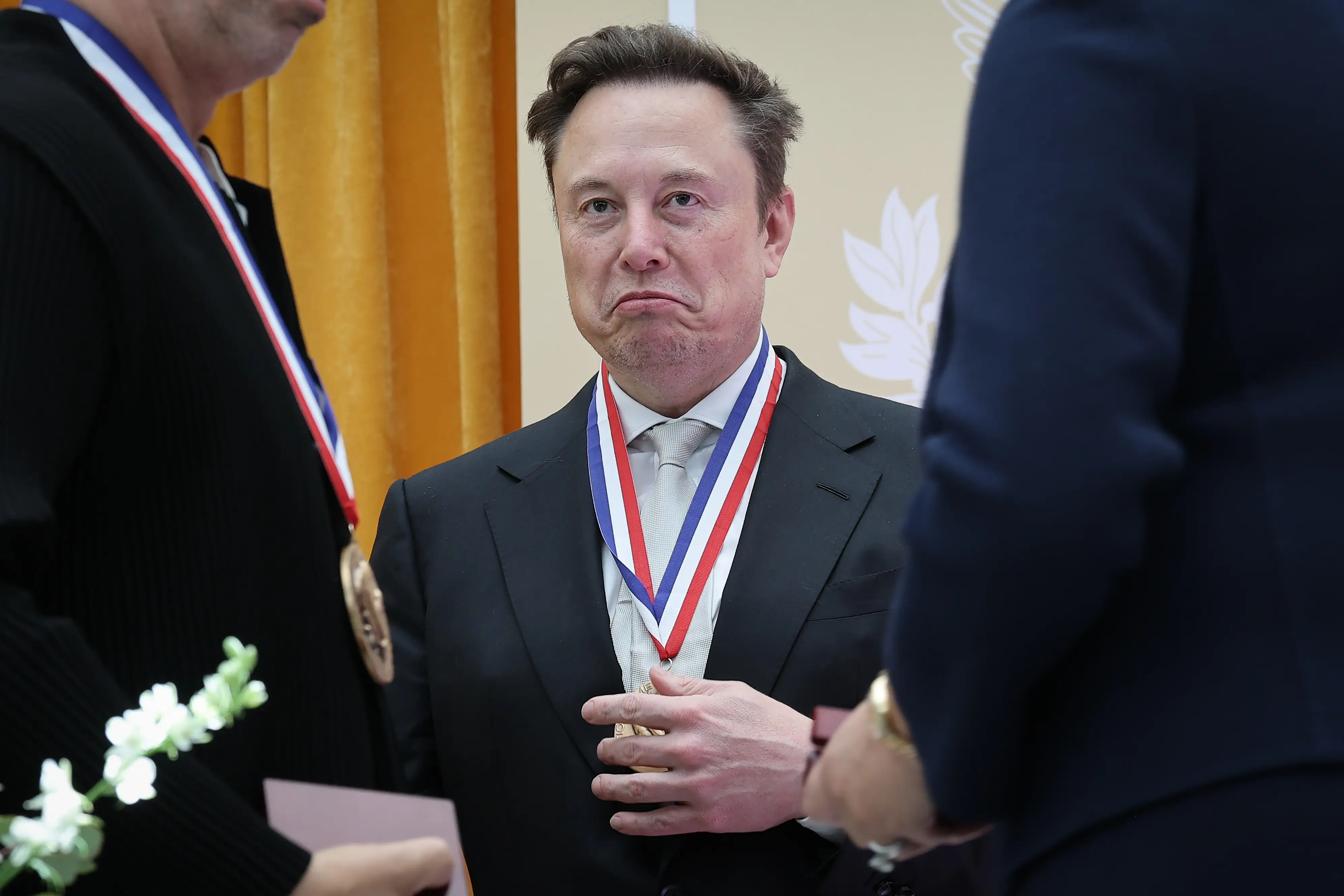 Elon Musk wears a medal on a ribbon while standing among formally dressed attendees at an indoor ceremony.