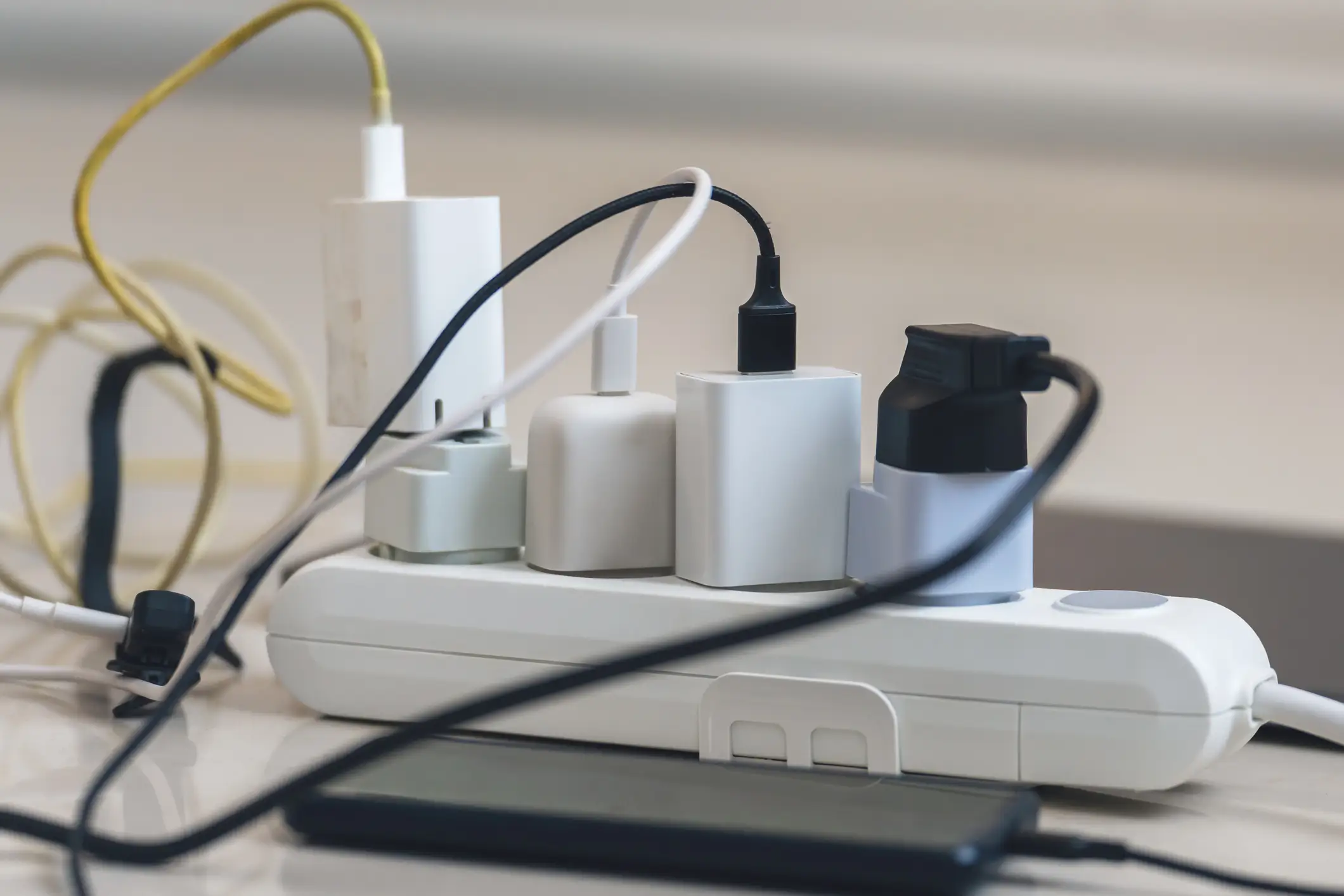 Multiple charging adapters and tangled cables are plugged into white power strips on a tabletop beside a smartphone.