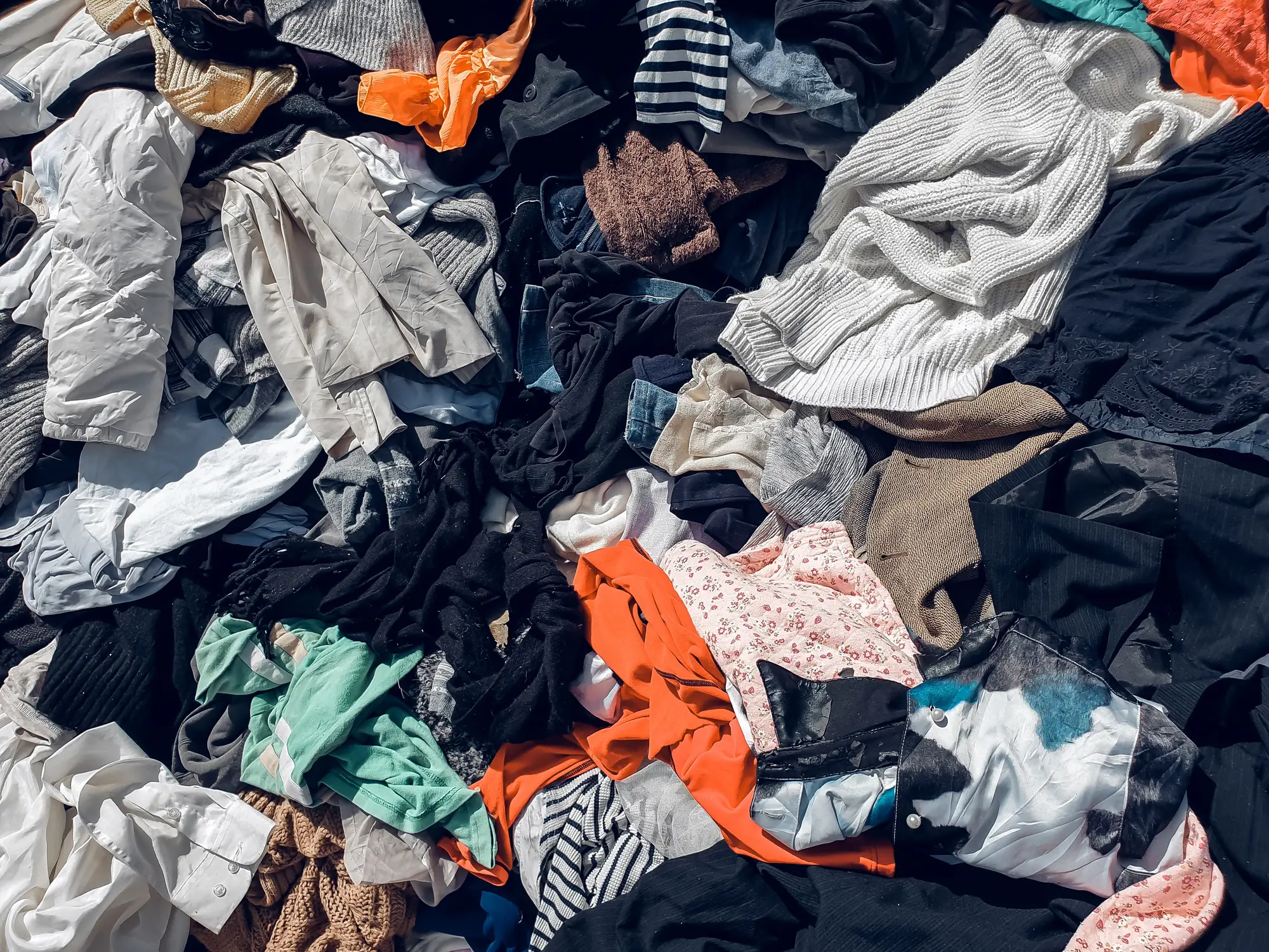 Assorted clothing items lie in a large sunlit pile, including sweaters, shirts, jackets, and patterned fabrics.
