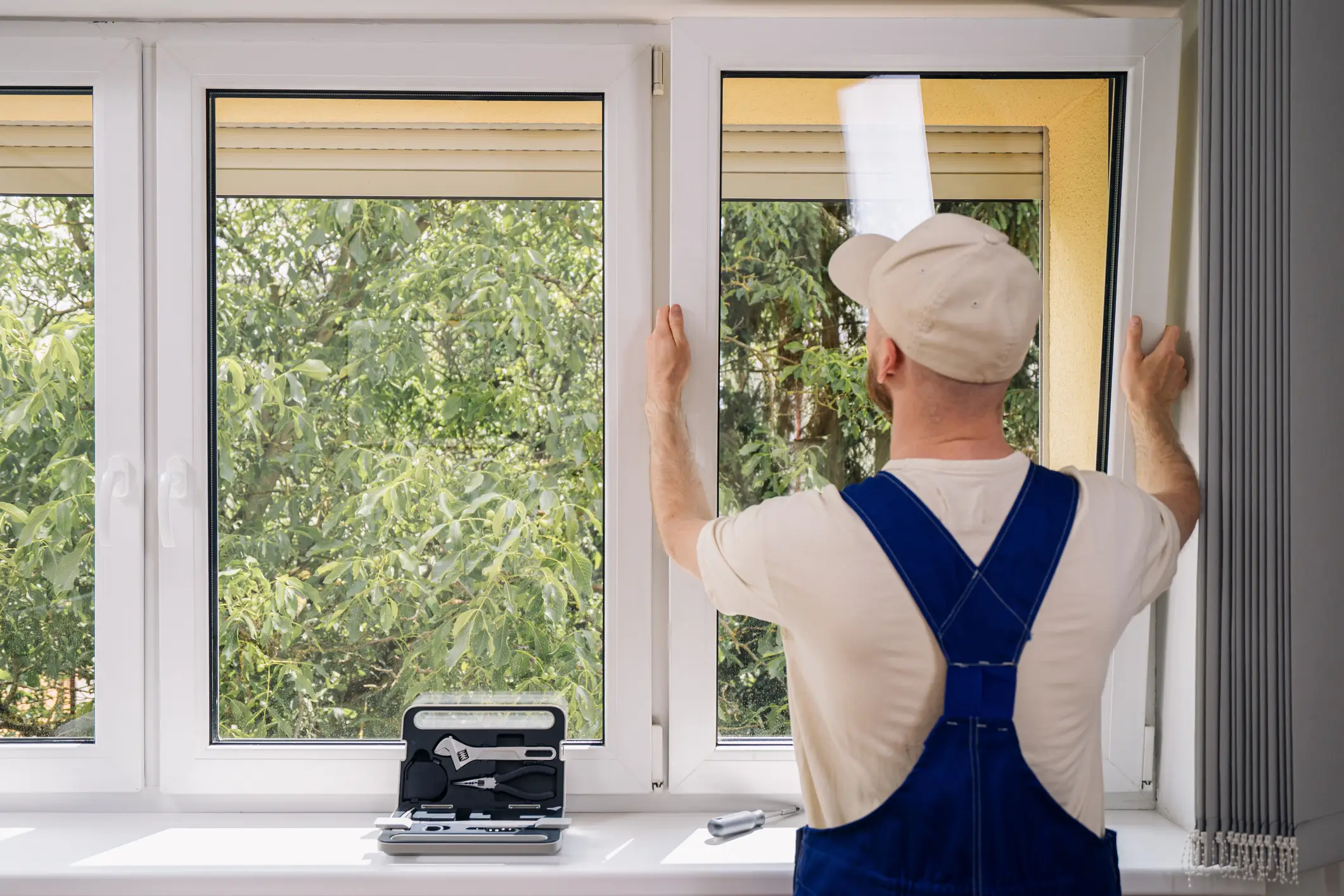 Worker in blue overalls adjusts a white interior window frame with a toolbox on the sill.